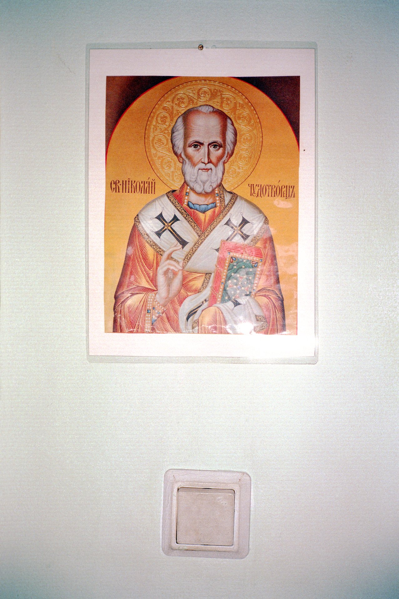 Orthodox Christian icon of Saint Nicholas in traditional robes with a halo, holding a book and making a blessing gesture, mounted above a wall switch.