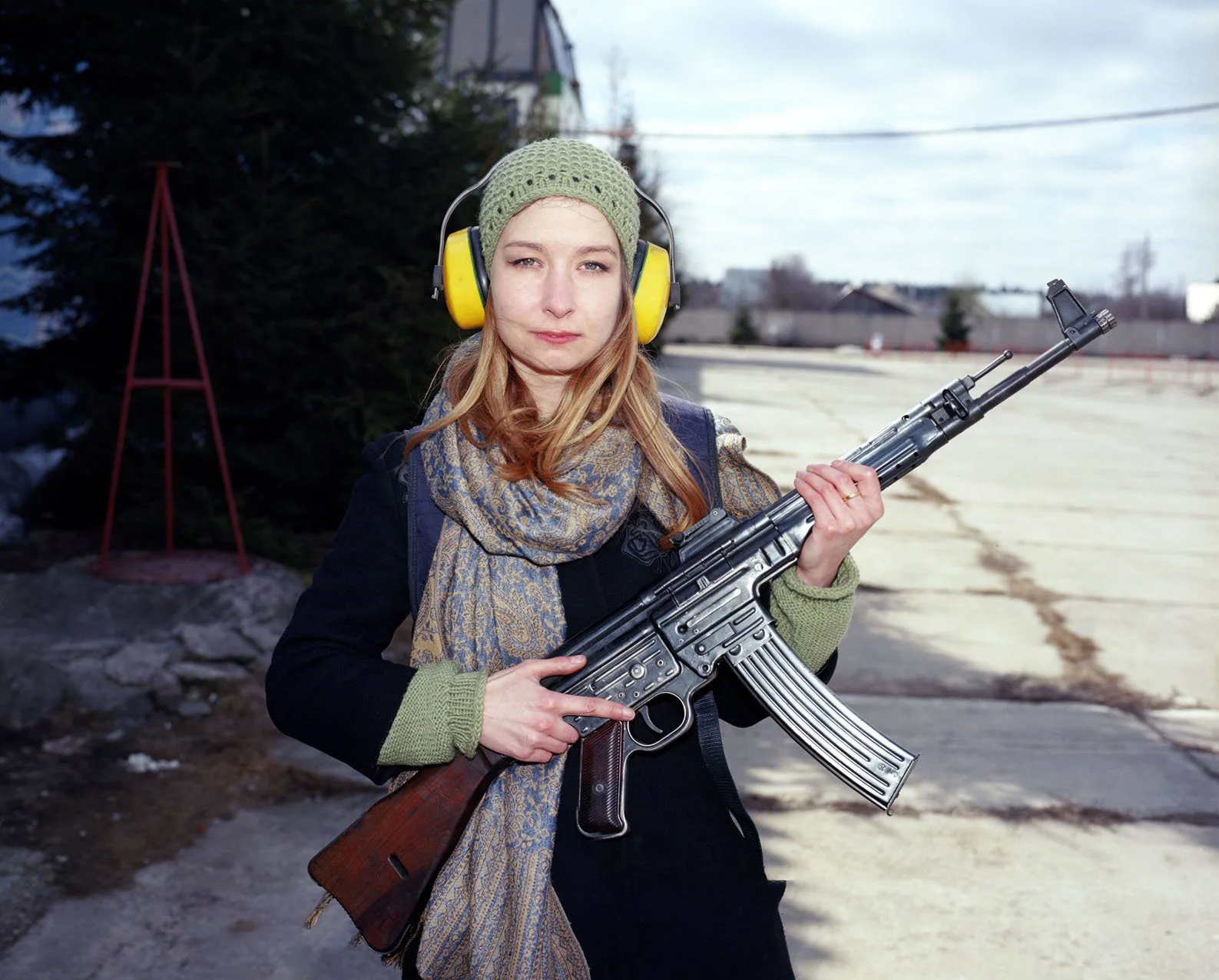 A woman with long hair wearing yellow ear protection, a green knitted hat, a scarf, a dark coat, and green gloves, holds a rifle outdoors with trees and a cloudy sky in the background.