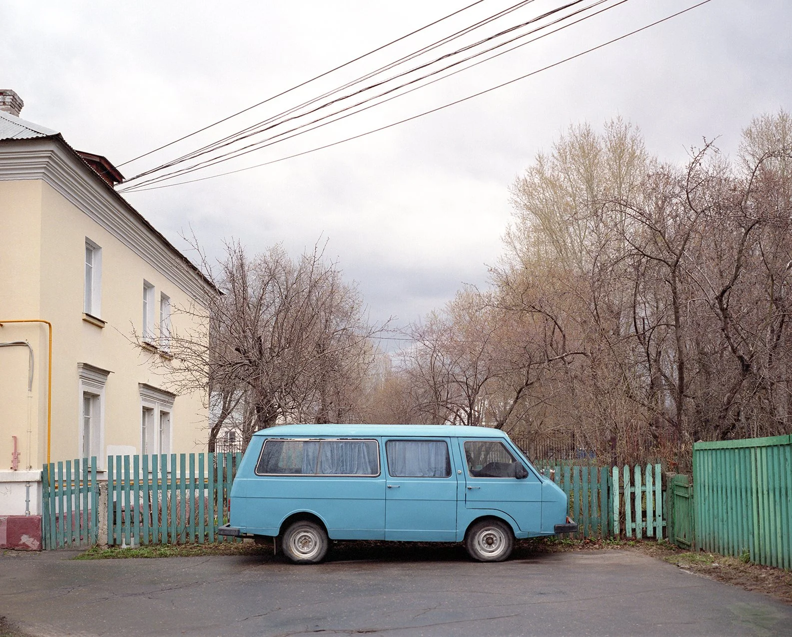 A vintage blue van parked on a street in front of a green wooden fence, with leafless trees and a pale yellow building with white windows in the background.