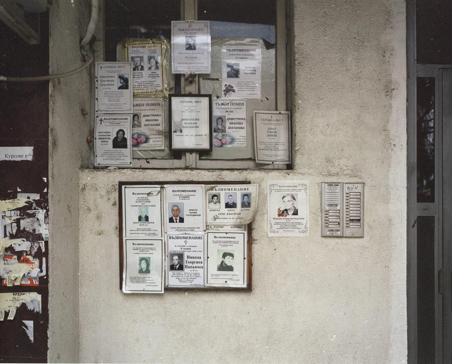An outdoor memorial display with funeral notices and photographs of deceased individuals, mounted on a concrete wall next to a glass door and a bulletin board with torn posters.