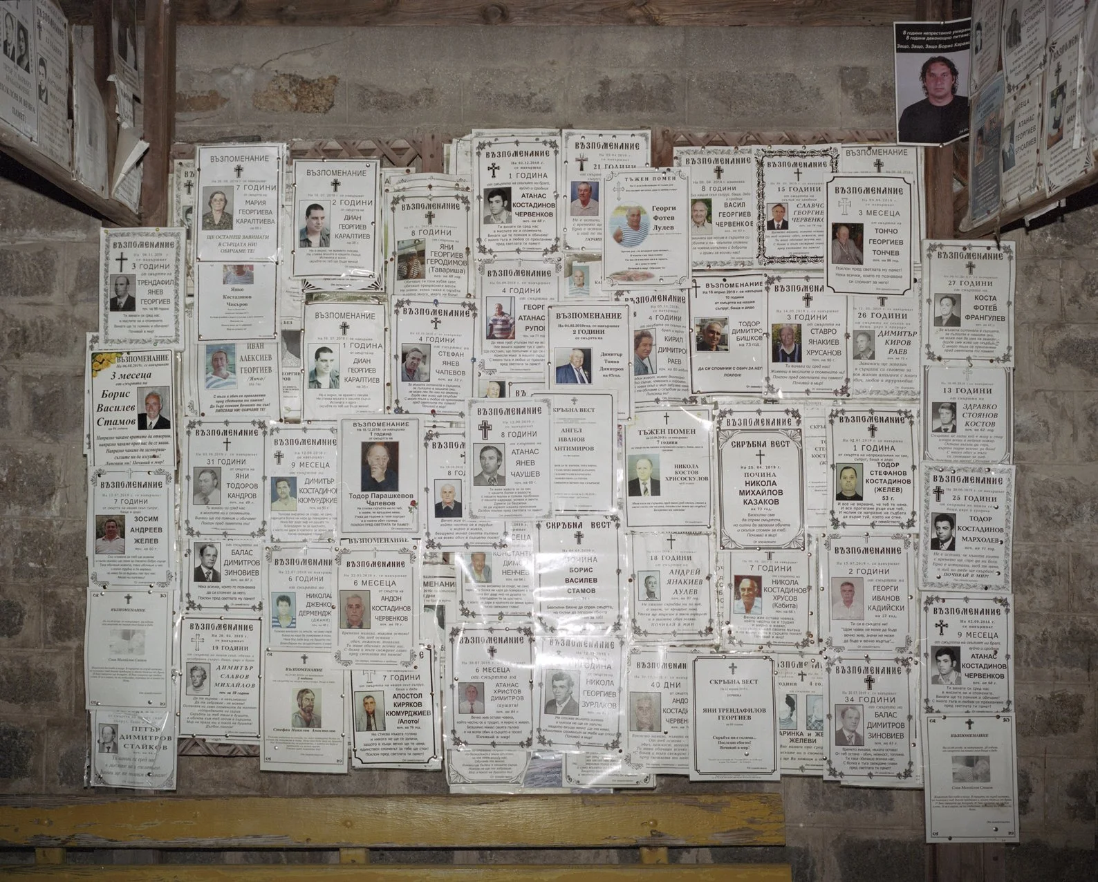 A memorial wall with many black-and-white obituary notices and photographs of deceased individuals, some with decorative borders and labels in a language using Cyrillic script, arranged on a wooden and brick background.