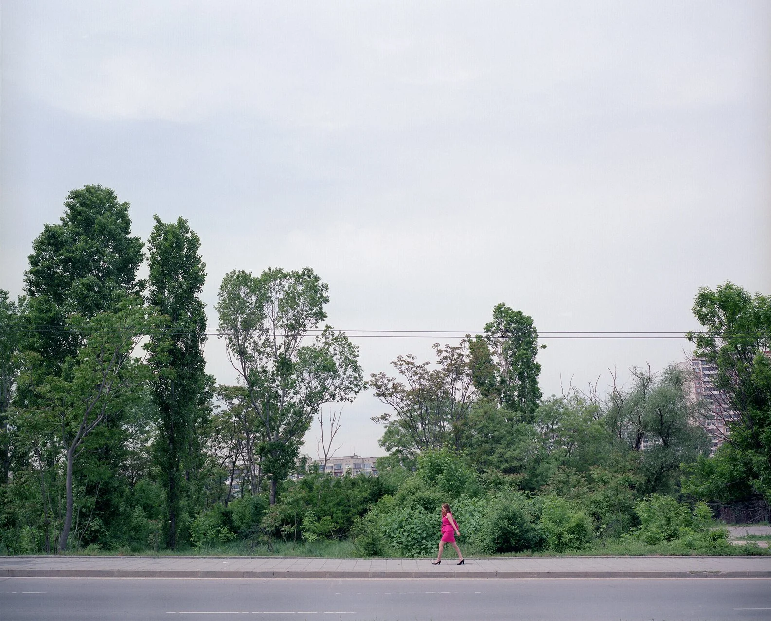 A woman in a pink dress walking along a sidewalk next to a green tree-filled area with an overcast sky.