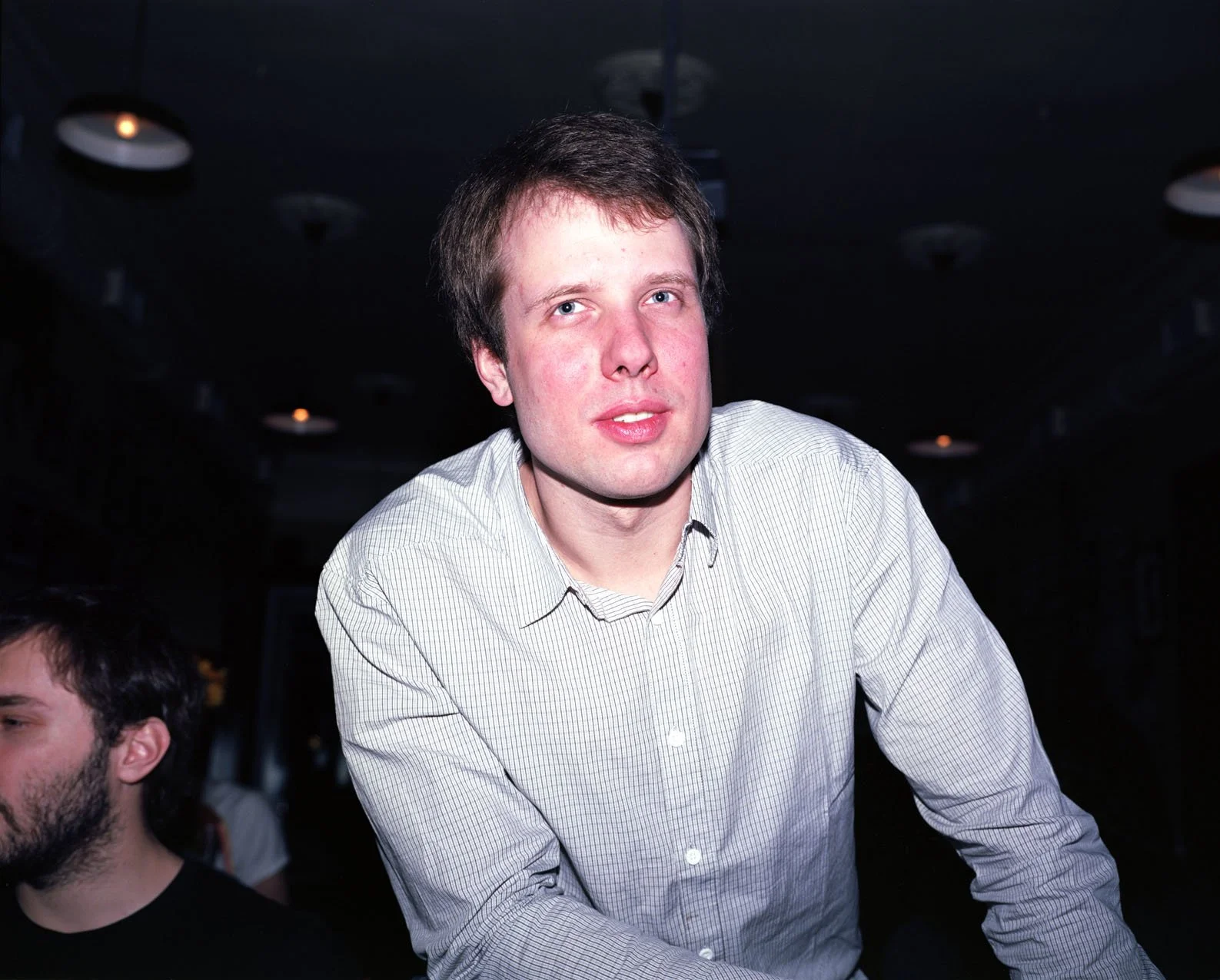 A young man with light skin, short brown hair, and blue eyes, wearing a light-colored, striped button-up shirt, looking at the camera with a neutral expression in a dark indoor setting.