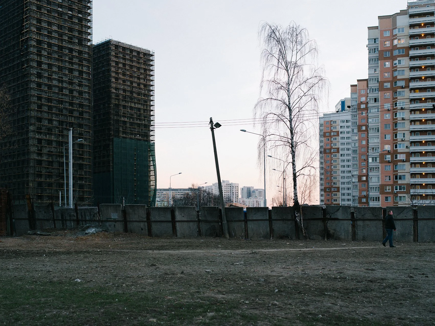 Cityscape with tall apartment buildings, a leafless tree, and construction site with temporary fencing, and a person walking along the sidewalk at dusk.