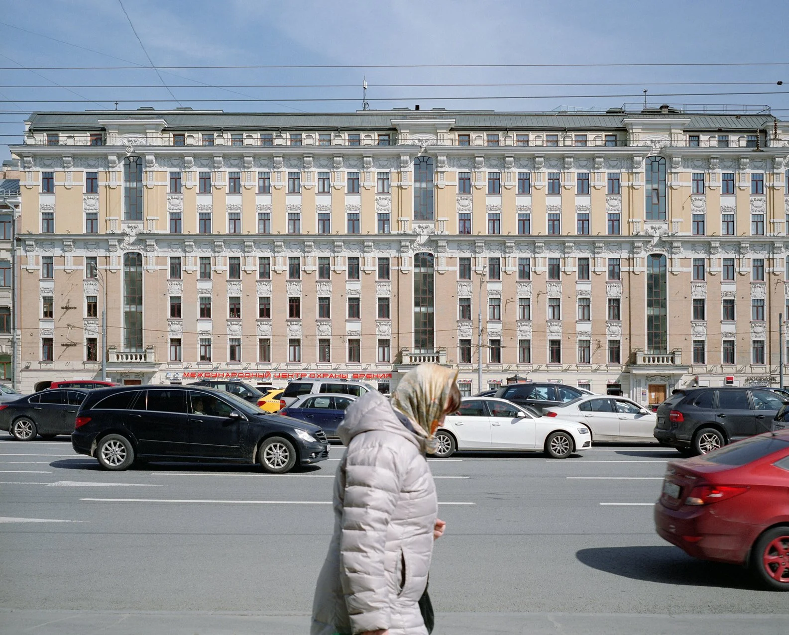 City street view with a woman in a white coat walking in front of a parking lot and a large, historic multi-story building in the background.