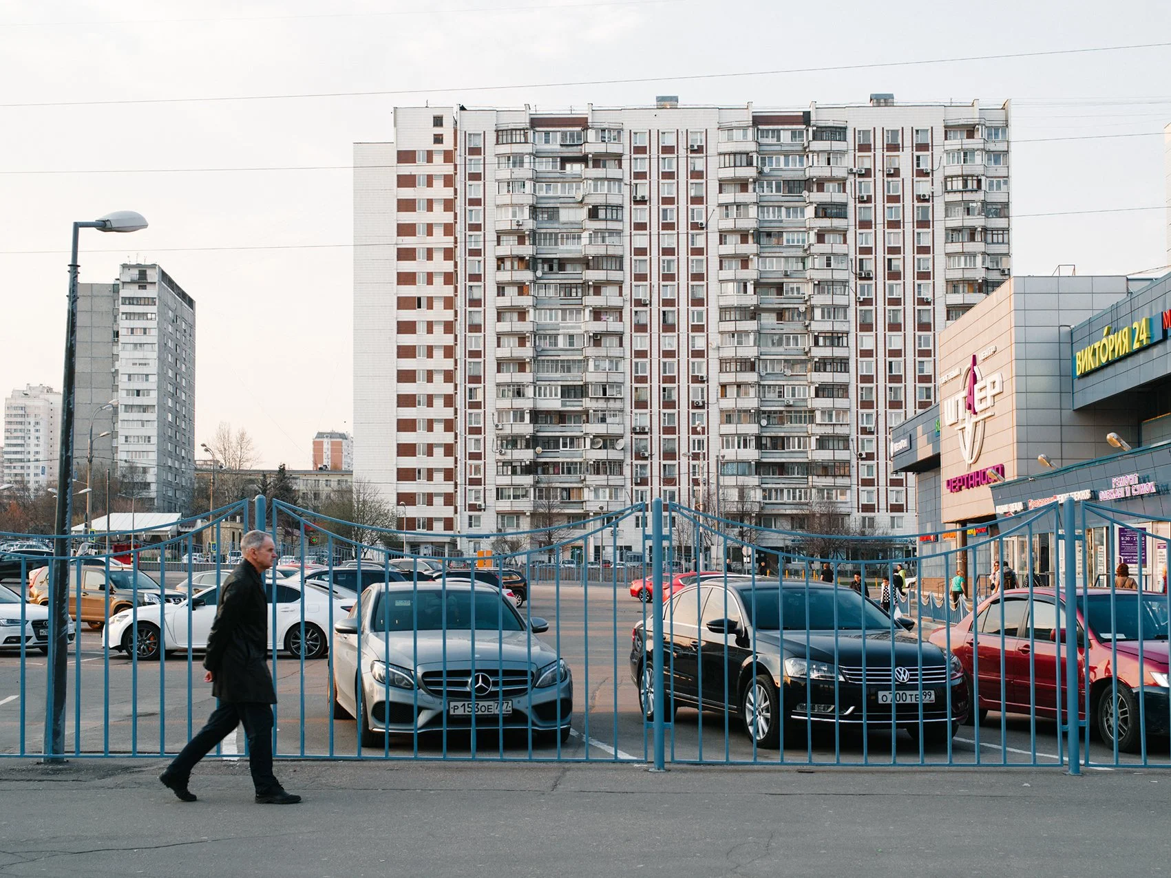 Urban scene with a parking lot filled with cars, a man walking past a blue gate, apartment buildings in the background, and a shopping center on the right.