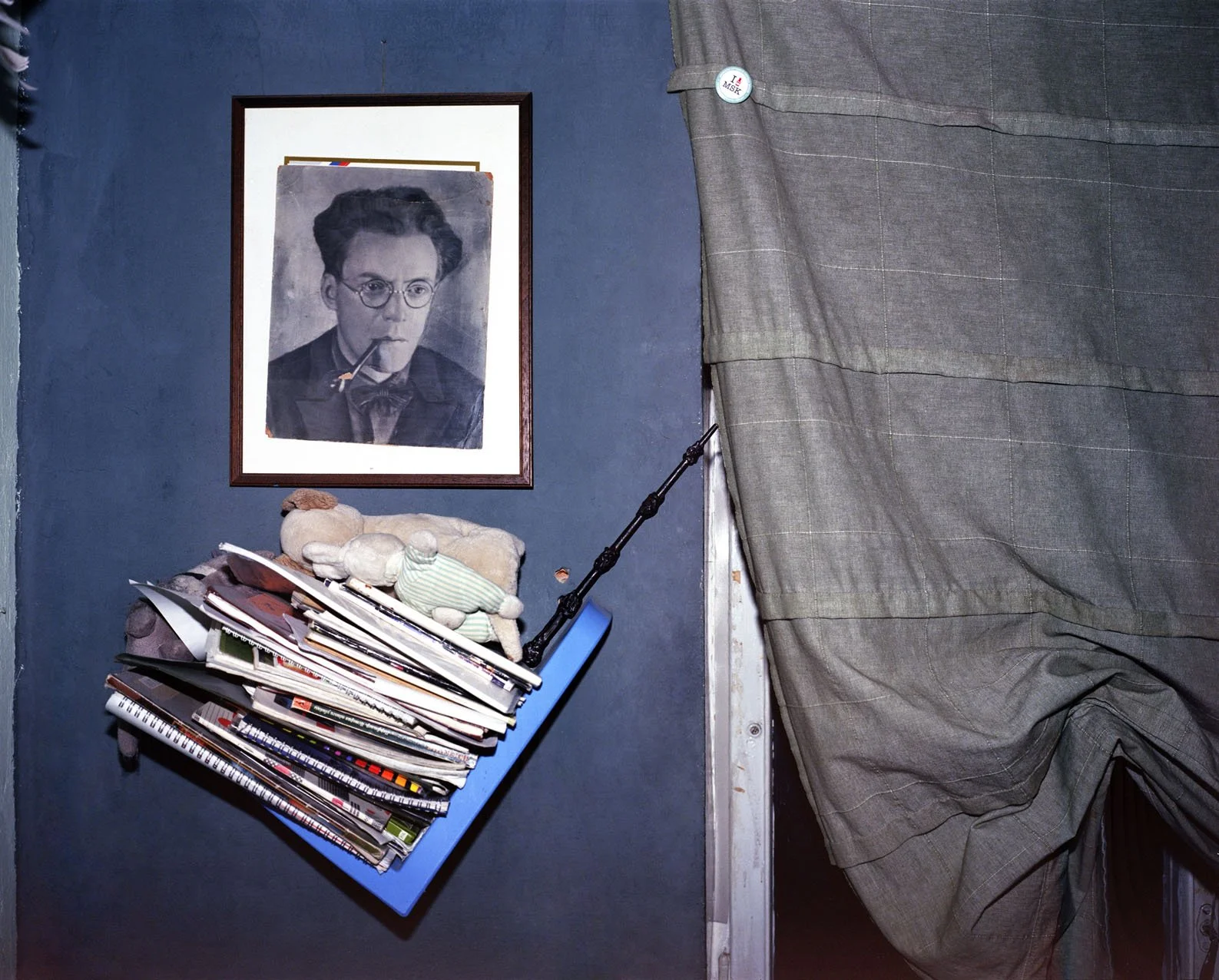 A wall with a framed black and white portrait of a woman wearing glasses and smoking a pipe, a pile of magazines and papers in a blue magazine holder, a stuffed toy bear with a green and white striped shirt, and a gray curtain partially covering a doorway.
