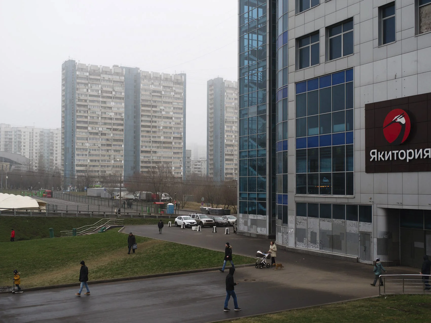 City scene with modern multi-story buildings, people walking on a wet pavement, some pushing strollers, and cars parked near a building with a sign that reads "Якитория".