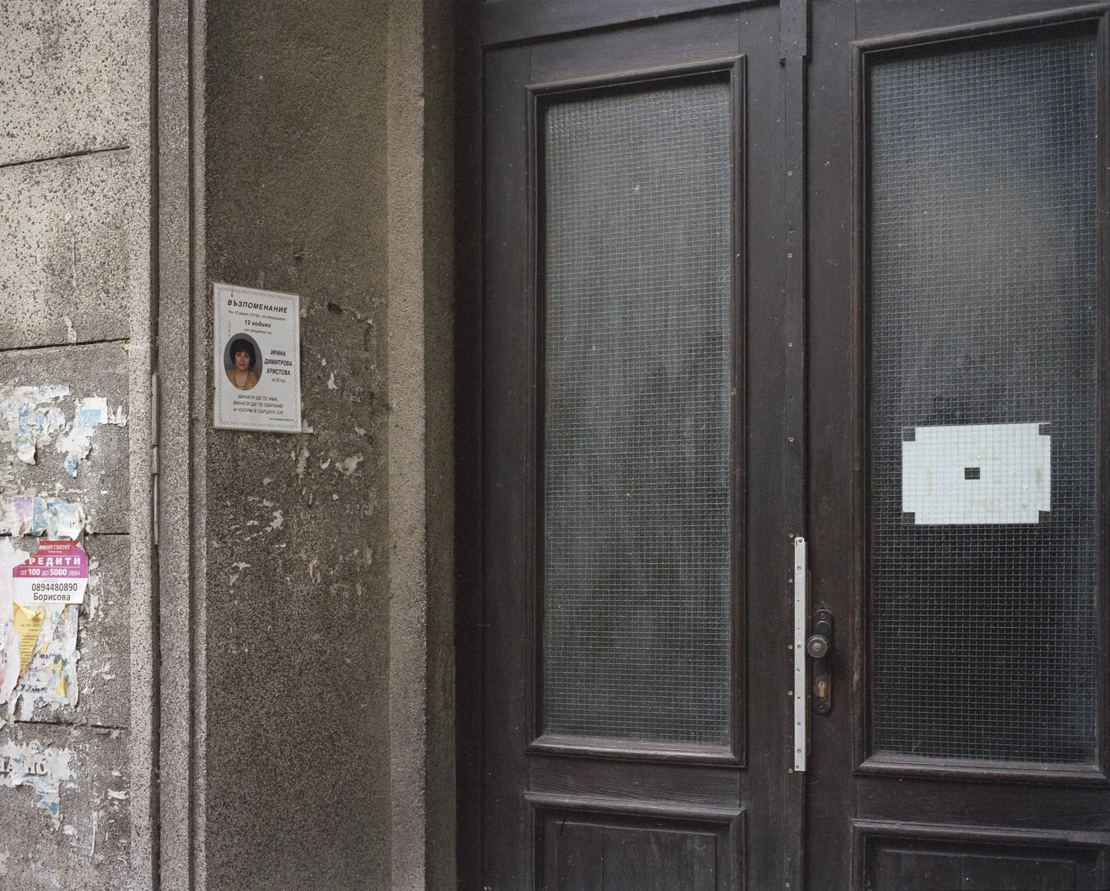 An exterior view of a dark brown door with glass panels, on a concrete wall with a memorial poster and some torn advertisements.