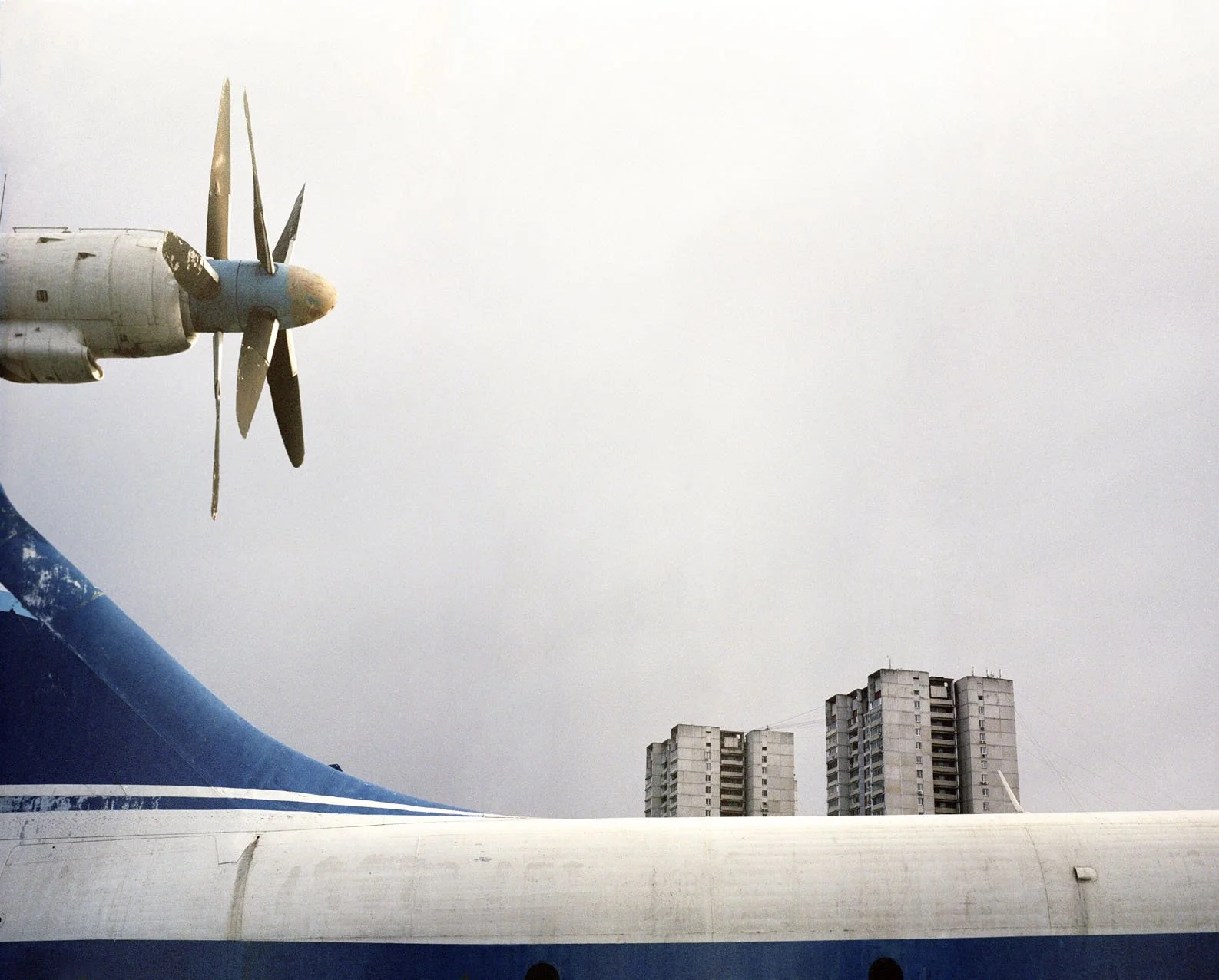Close-up of a large airplane's wing with a visible propeller against a cloudy sky, with tall residential buildings visible in the background.