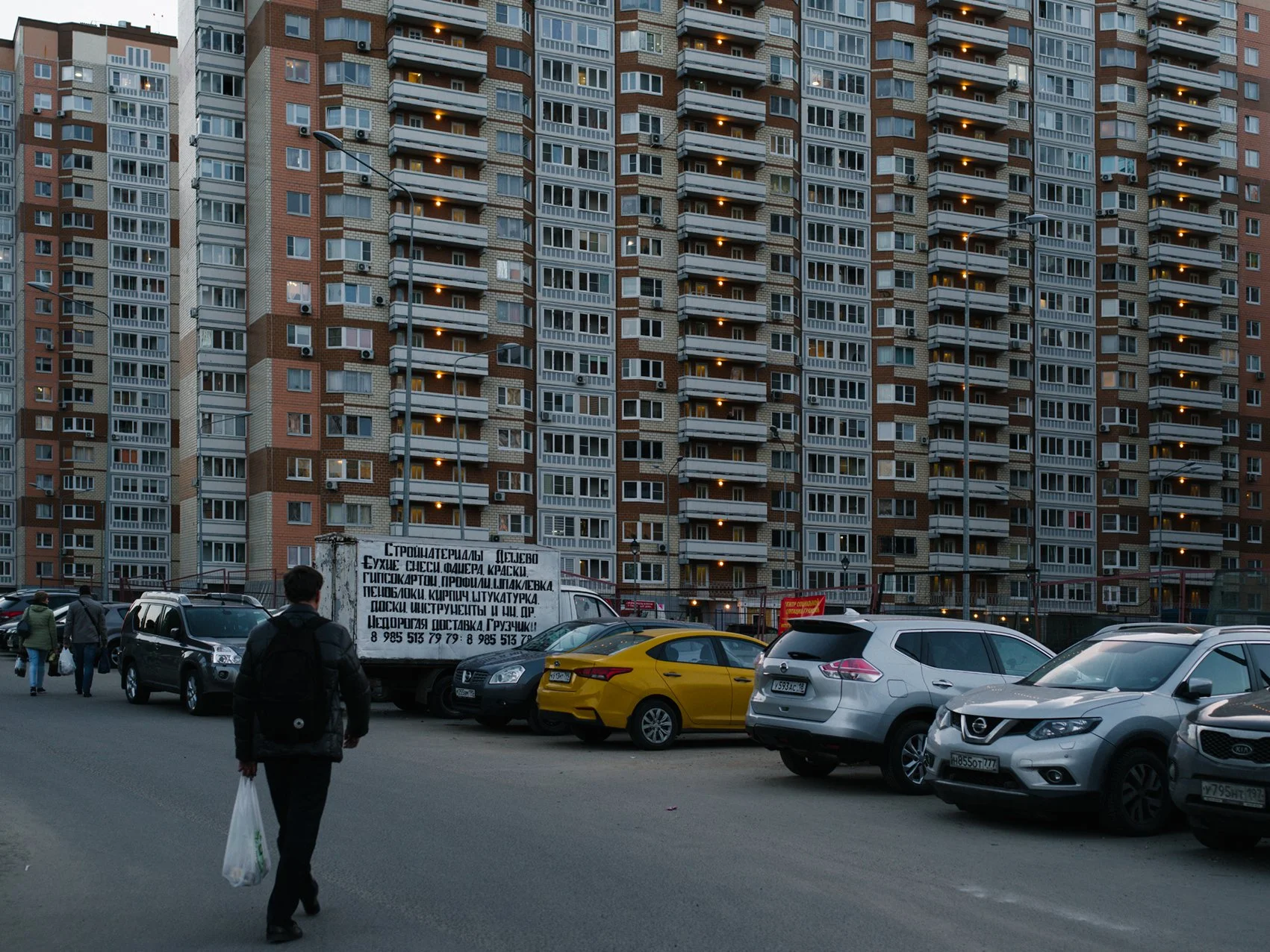 People walking on the street with parked cars in front of a tall apartment building during dusk.