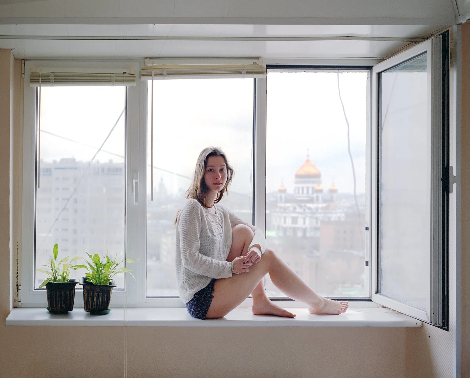 A young woman sitting on a windowsill with an urban cityscape and a domed building in the background, next to two potted plants.