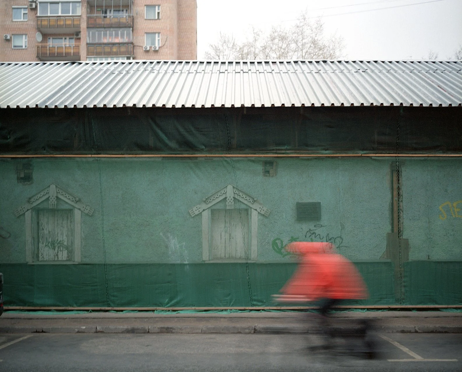 Blurred person wearing an orange raincoat walking past a green wall with graffiti and two small house-shaped windows, in an urban setting with a corrugated metal roof and a multi-story brick building in the background.