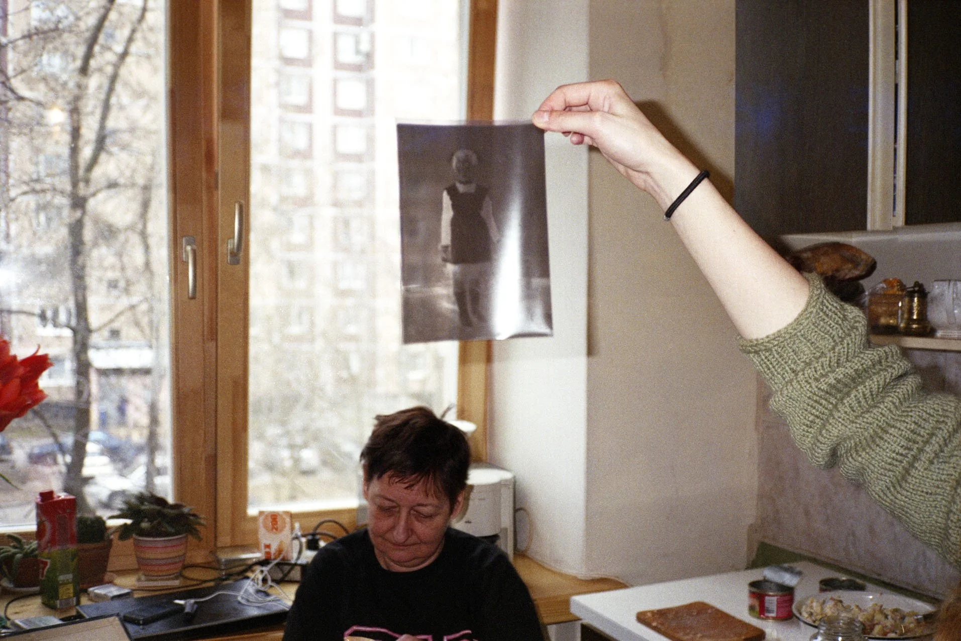 Person holding up a black-and-white photograph of a woman in front of a window with a cityscape outside. A woman with short dark hair is sitting at a table nearby.