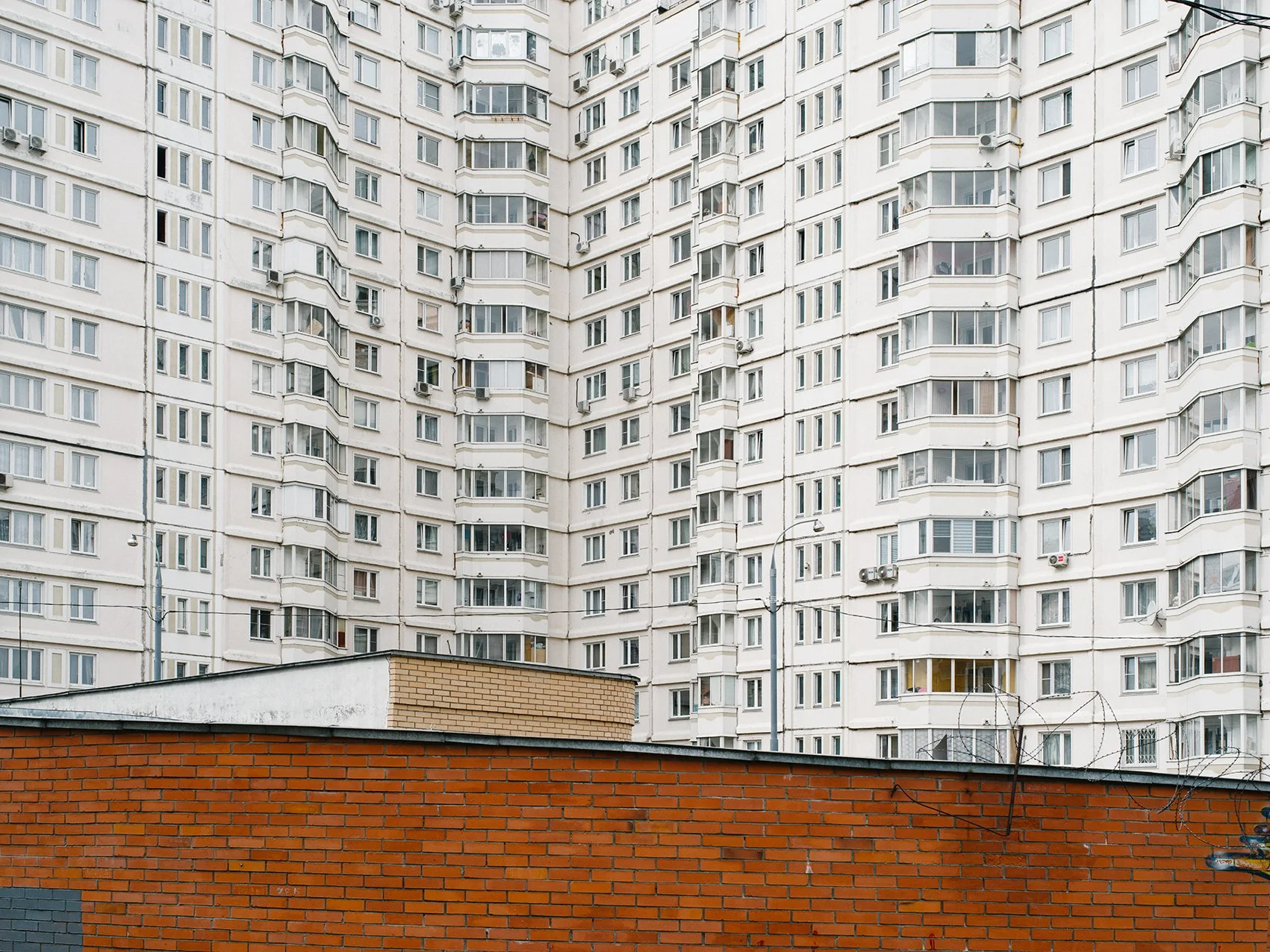 A tall, white, multi-story apartment building with numerous windows and balconies, seen behind a red brick wall and a small brick structure.