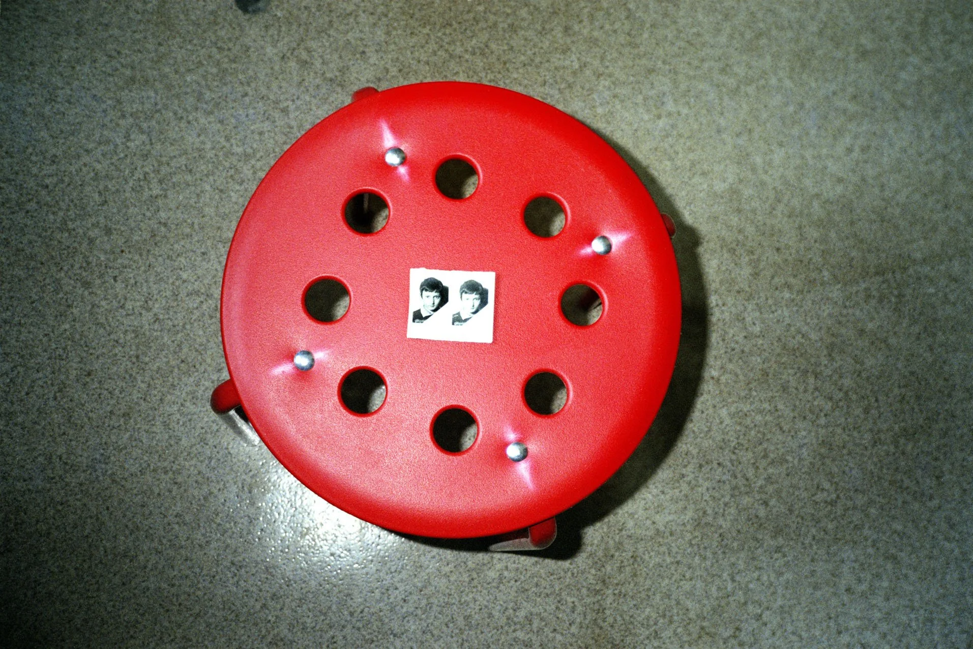 A red spaghetti strainer with six holes and four small metallic balls placed on top, and a small photo of a woman with short hair in the center.