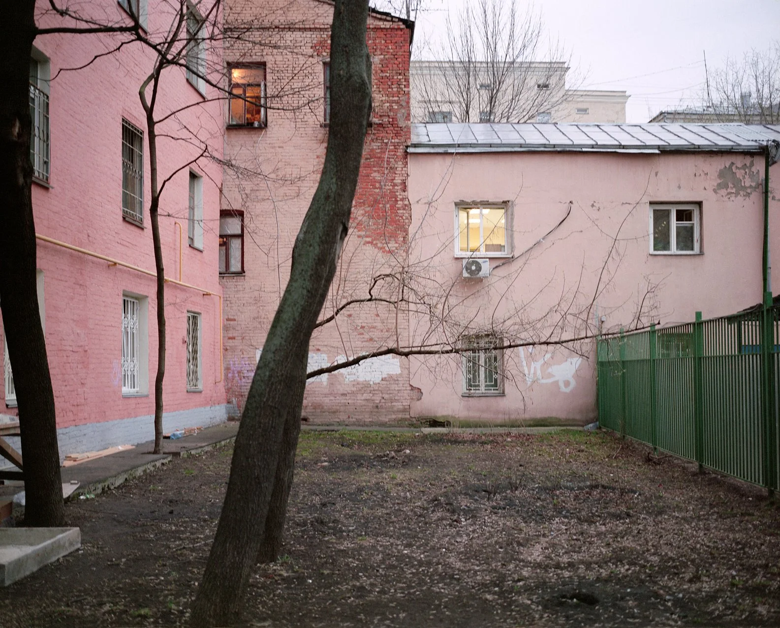 A view of an urban backyard with a pink and red brick building, leafless trees, and a green metal fence, during dusk.