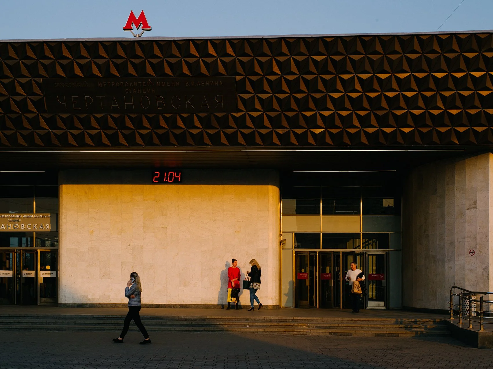 People entering and exiting the Chertanovskaya metro station in Moscow, Russia, with a digital clock displaying 21:04, and an architectural facade with a geometric pattern.