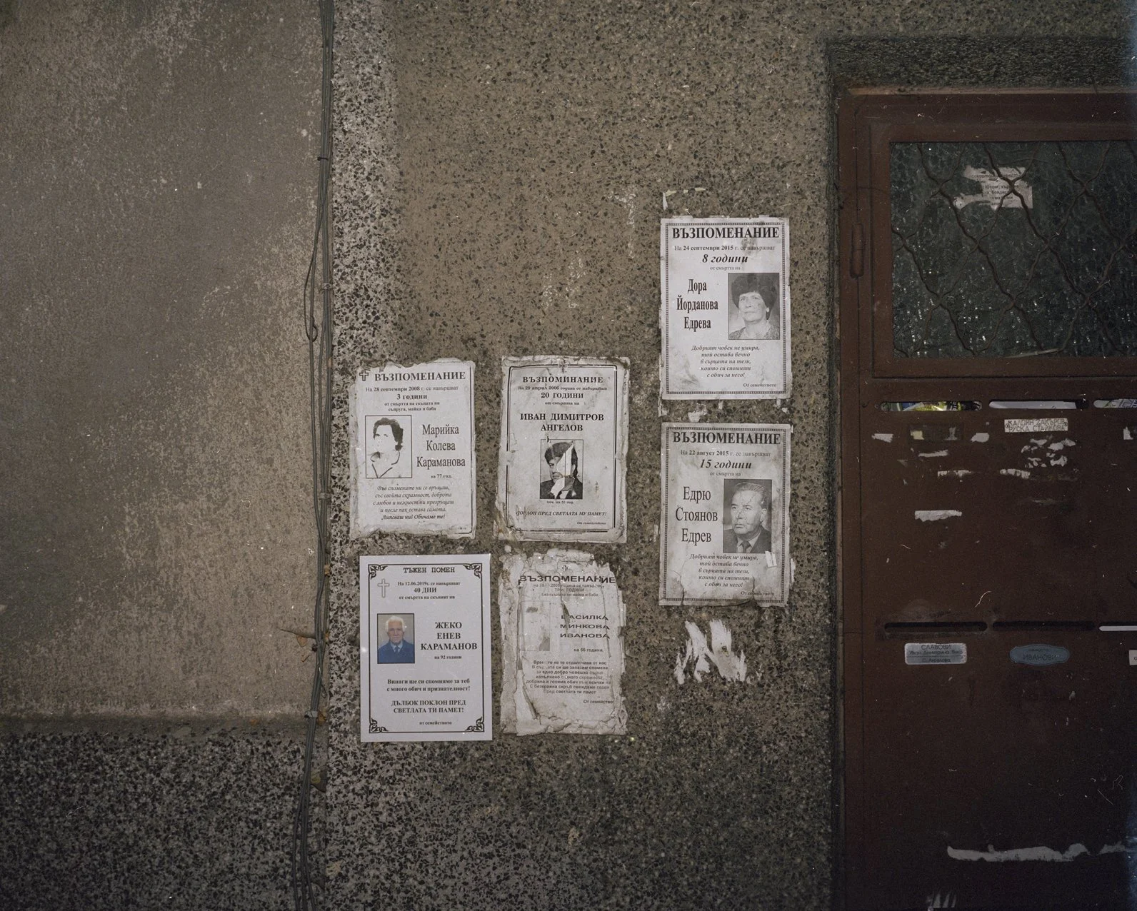 Memorial notices for multiple individuals on a concrete wall near a brown door, including photos, dates, and messages in Bulgarian.