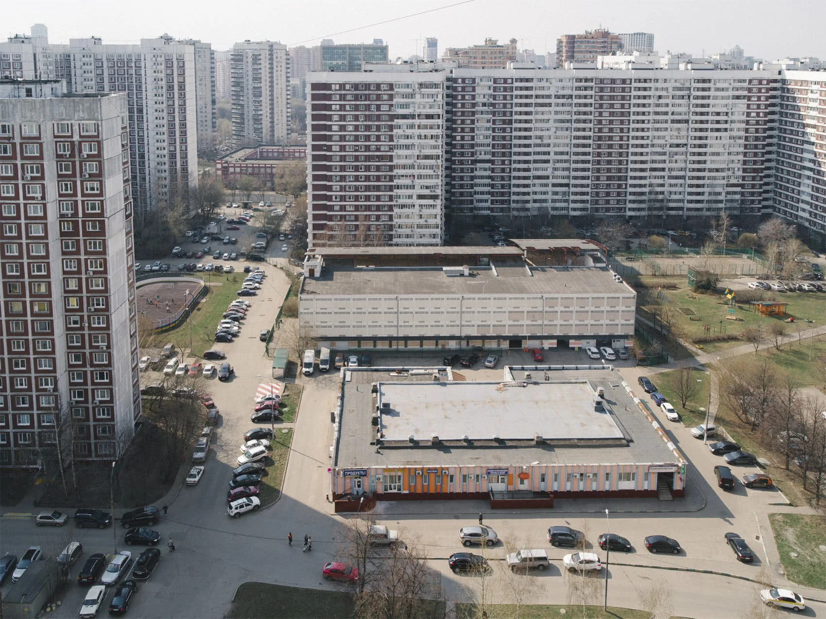 Aerial view of a city with tall residential buildings, parking lots, and a small commercial building in the foreground, alongside green spaces and playgrounds.