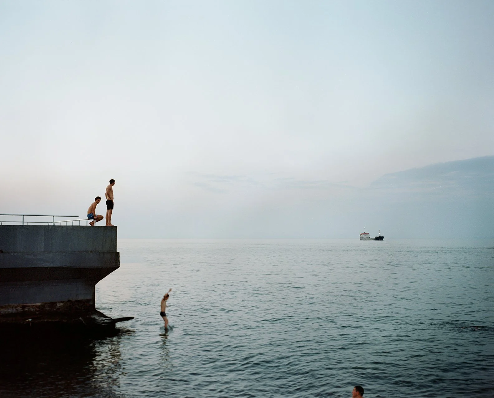 Three young men on a concrete pier with one jumping into the water and two others preparing to jump, with a ship in the distance on the ocean under a calm, cloudy sky.