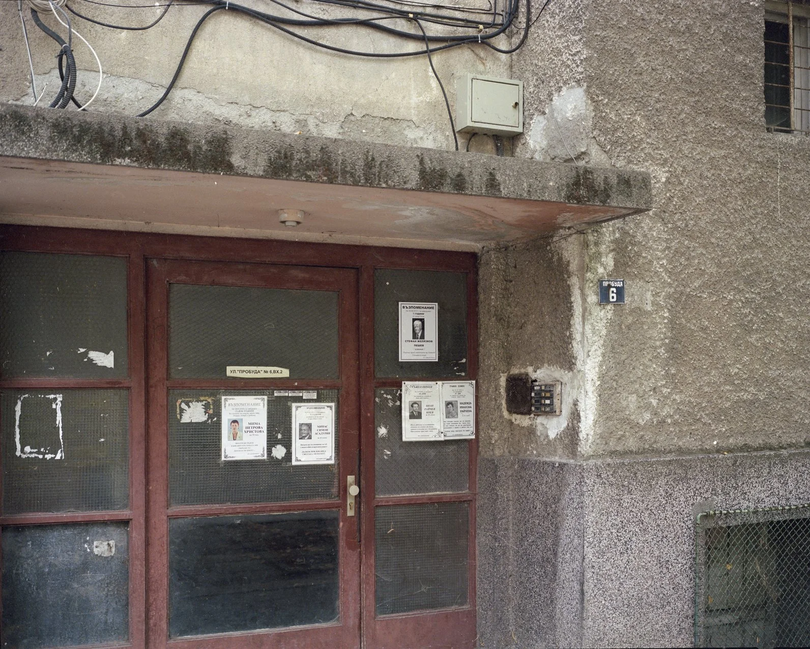 Entrance to an old building with cracked concrete walls and a red wooden door with glass panels. Several notices and flyers are posted on the door. There is a small metal box, a street sign with the number 6, and a keypad lock near the door. Electrical wires run along the upper part of the building.