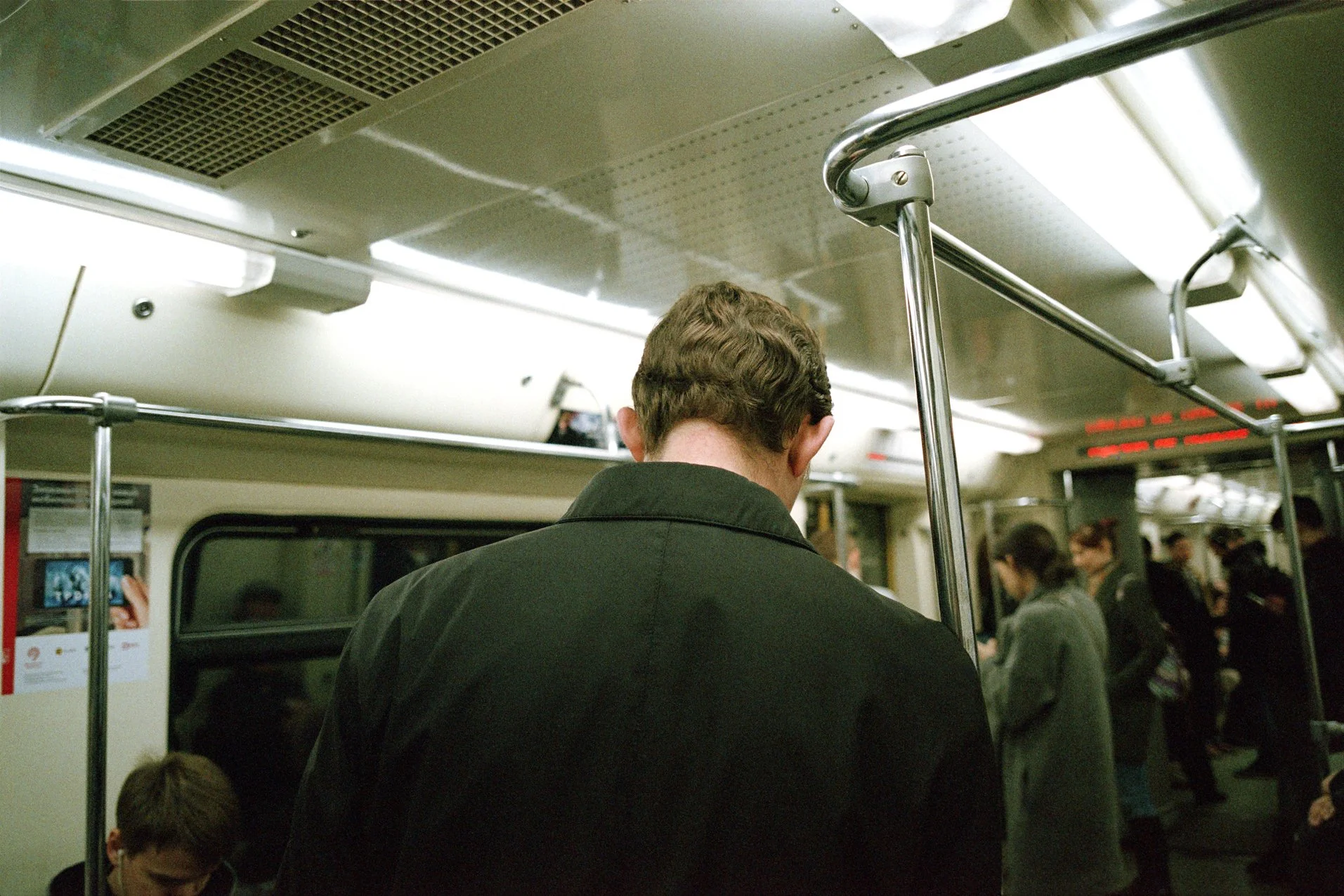 Back view of a man with brown curly hair wearing a black jacket standing inside a subway train, with other passengers in the background.