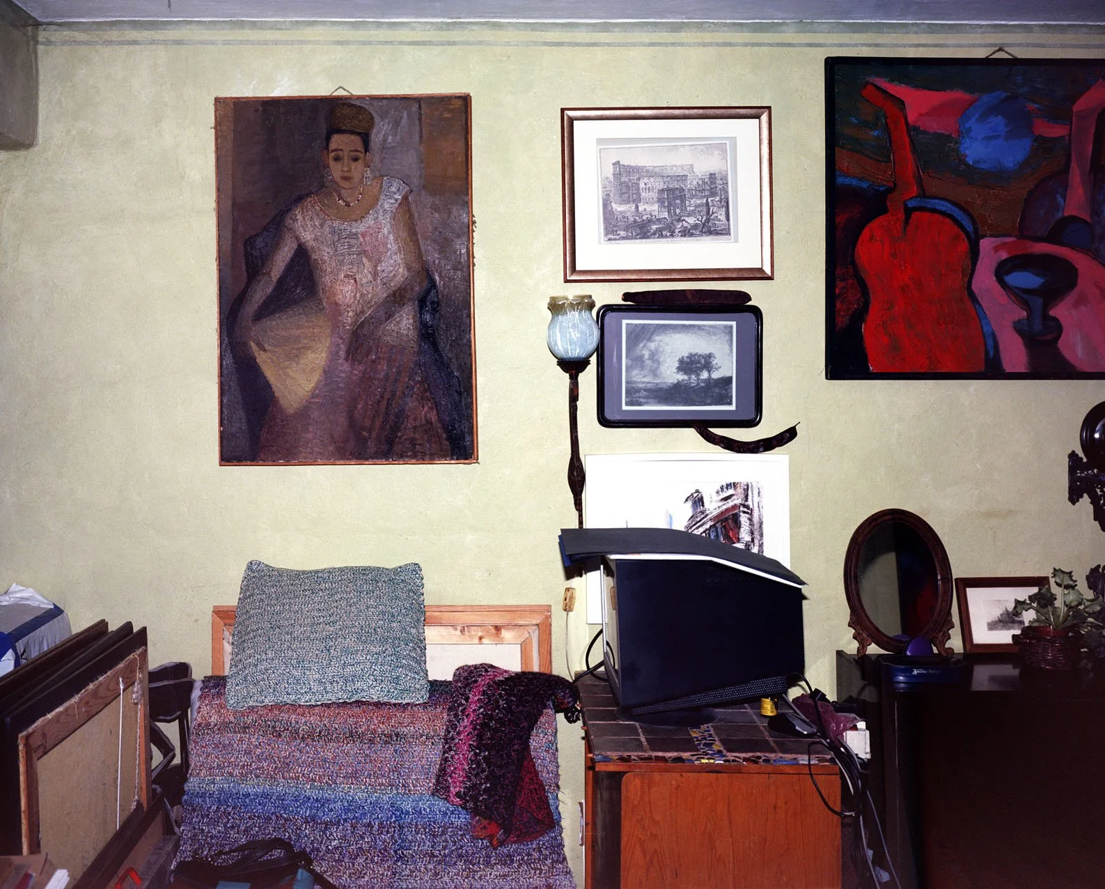 A cluttered room with various framed artworks on a yellow wall, including portraits and landscapes, a patterned armchair with a pillow and blanket, and a desk with a computer monitor, a mirror, and decorative items.