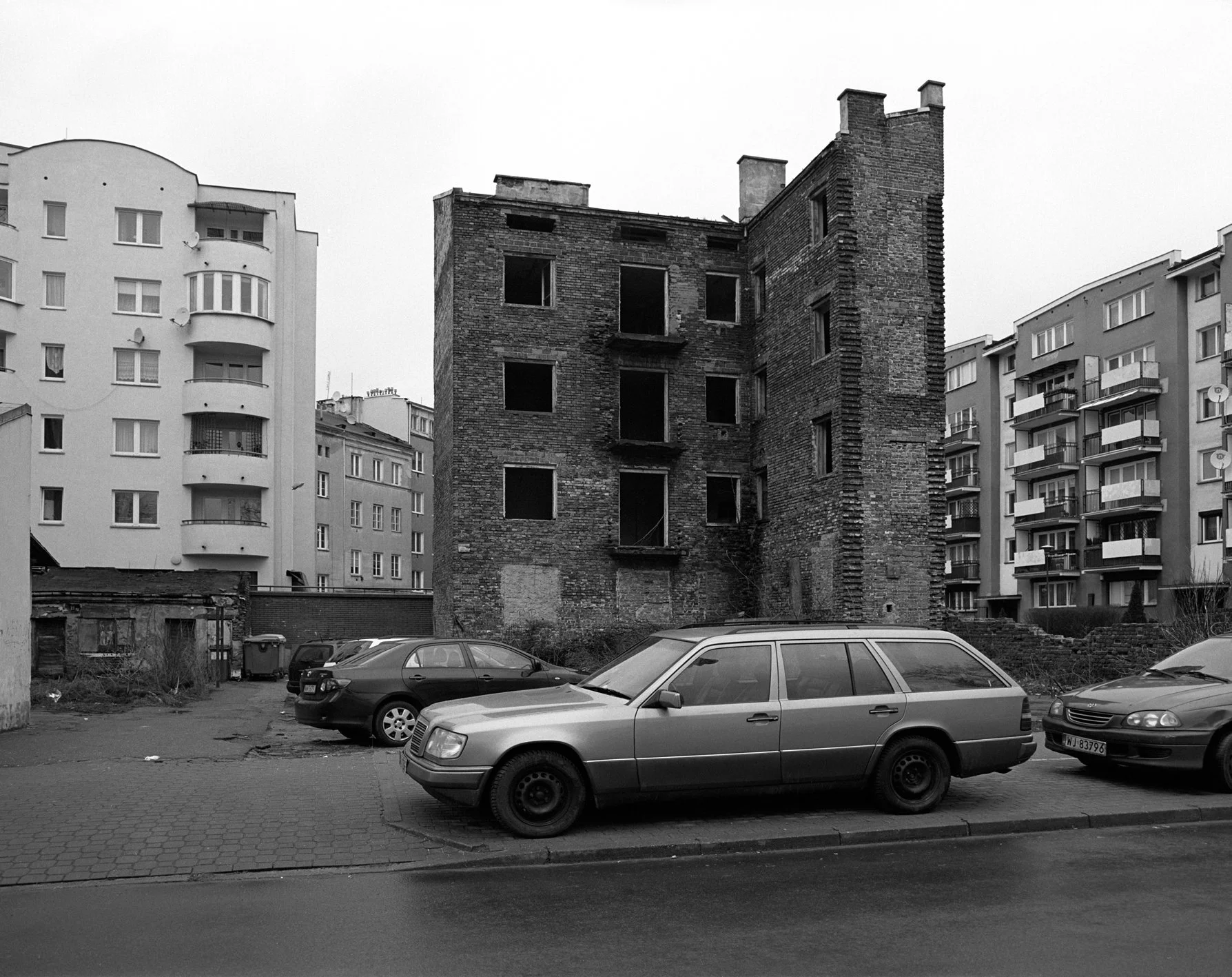 An urban scene with parked cars in front of an abandoned brick building and modern apartment buildings in the background.