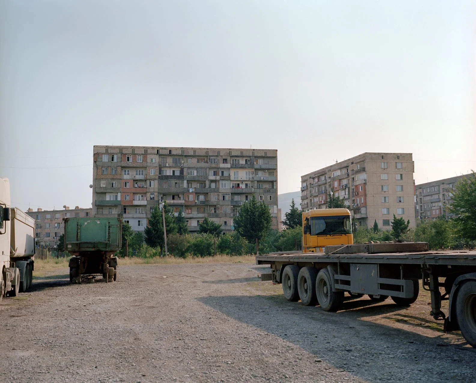 Empty gravel lot with parked trucks and a school bus, with apartment buildings and trees in the background.