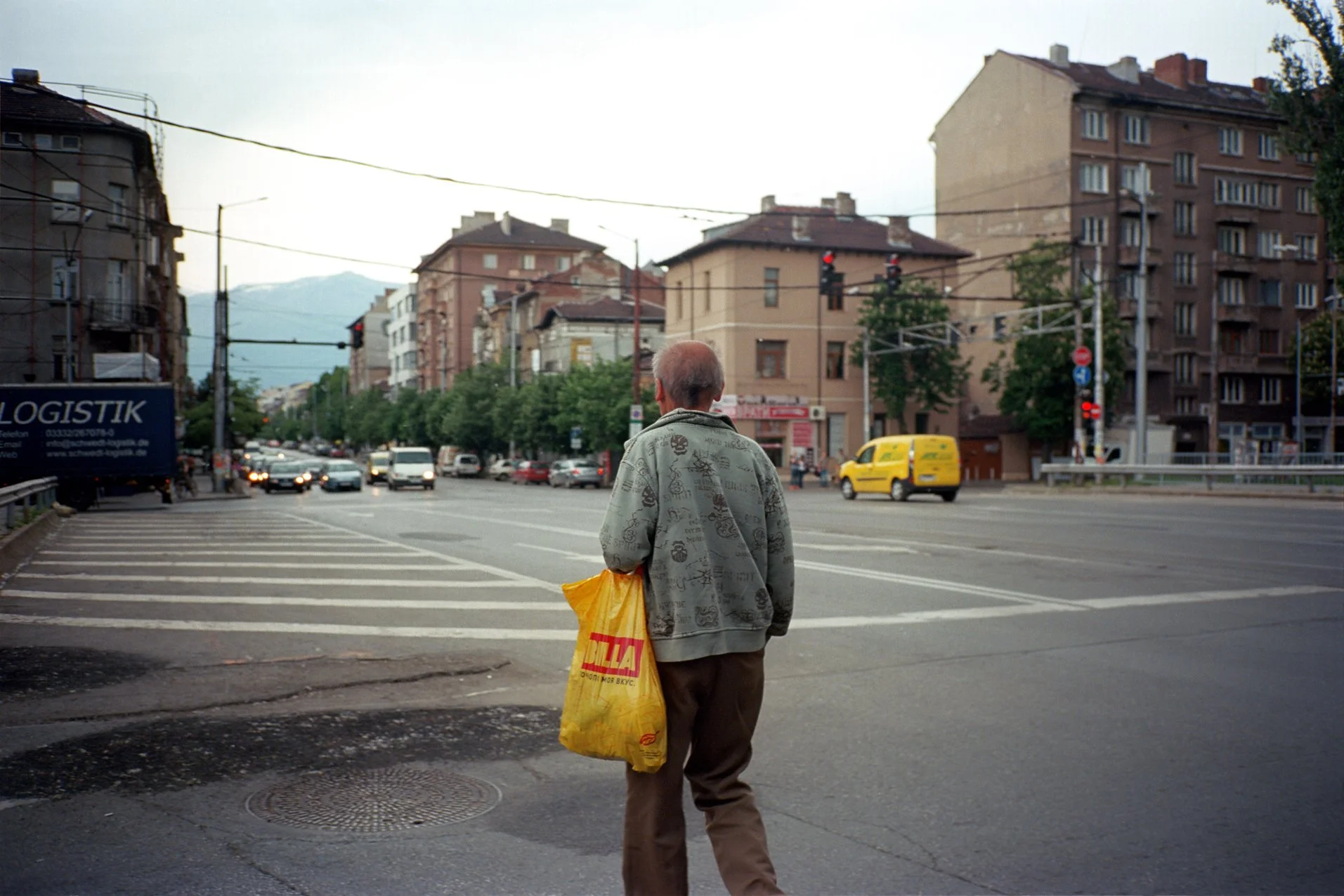 An elderly man walks across a busy city street carrying a yellow plastic bag. The background features several buildings, vehicles, and traffic signals.