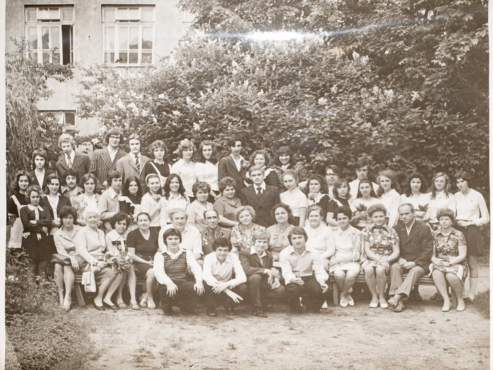 A group of people, including children, teenagers, and adults, gathered outdoors in front of a garden with trees and bushes for a photo.