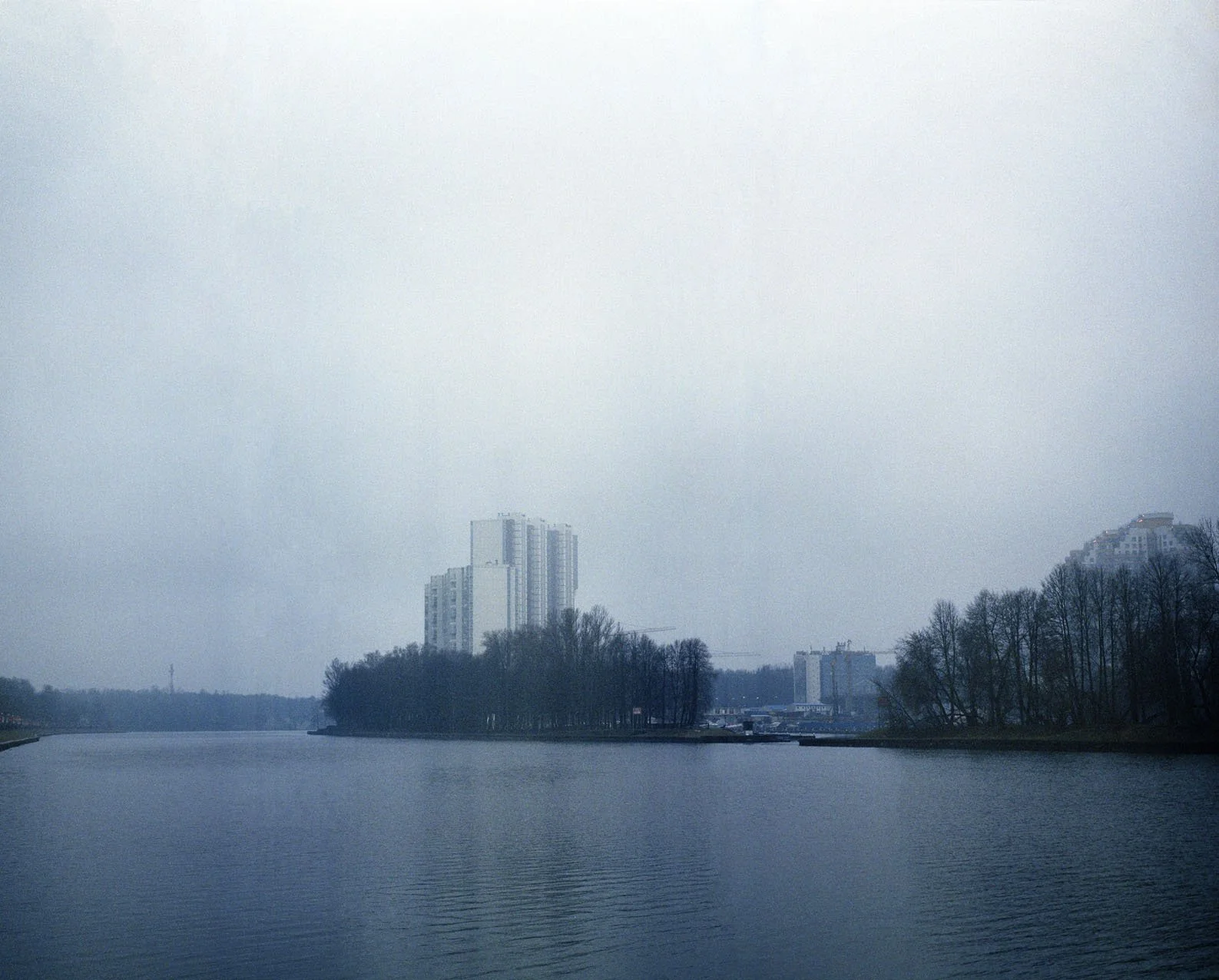 A body of water with trees along the shoreline and tall buildings in the background, under an overcast sky.