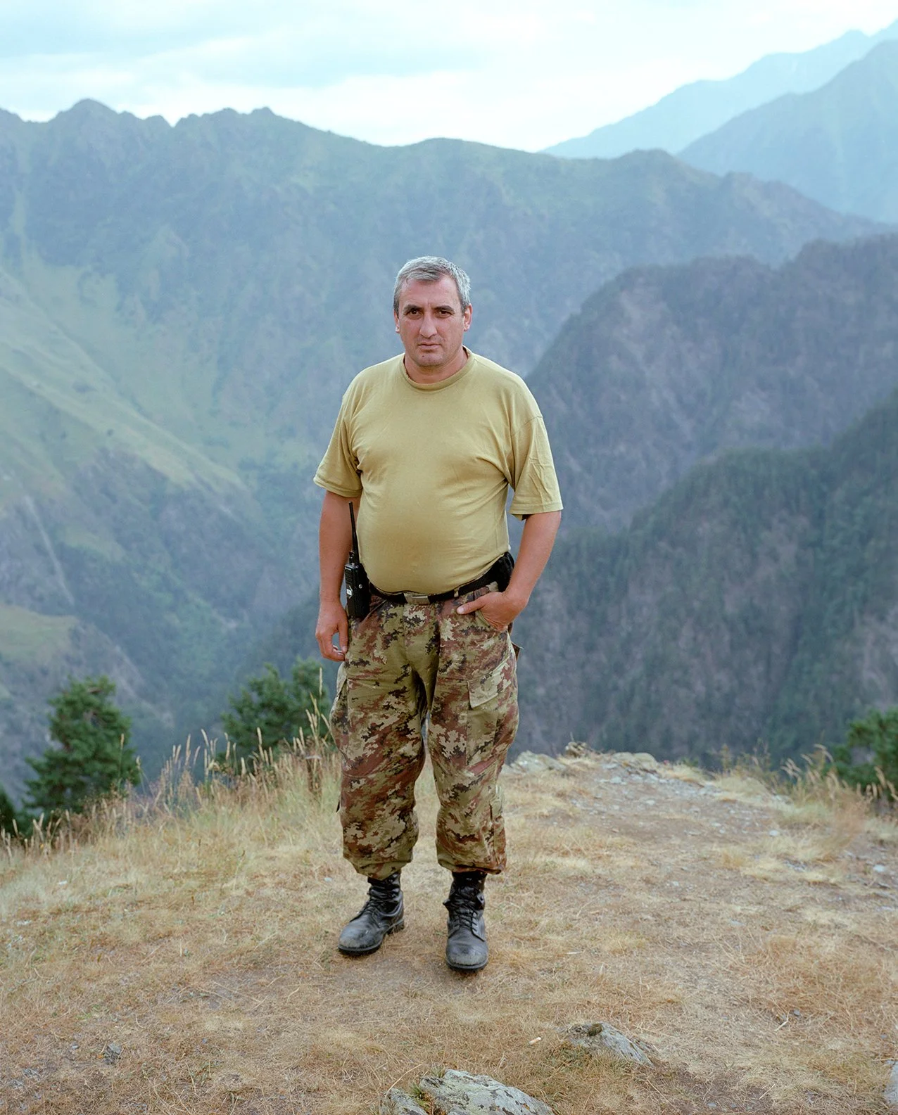 A man stands on a grassy hill overlooking mountainous terrain, wearing military camouflage pants, a yellow t-shirt, and black boots. He has a walkie-talkie on his belt and has his hand in his pocket.