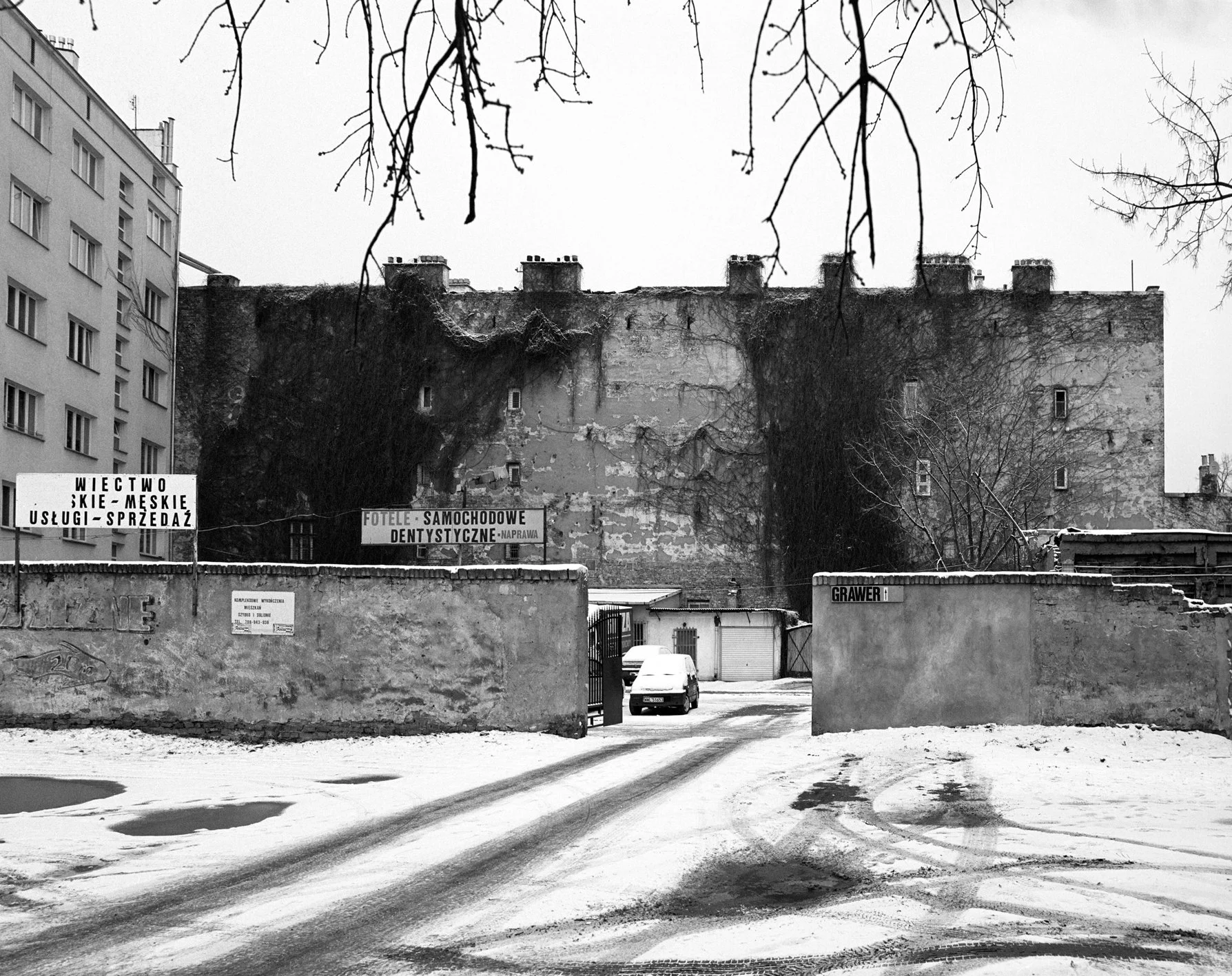 A black and white photo of a snowy, icy street entrance to an alley with a tall brick wall and an older building with ivy covering parts of it in the background. There are signs in Polish and a few cars parked.