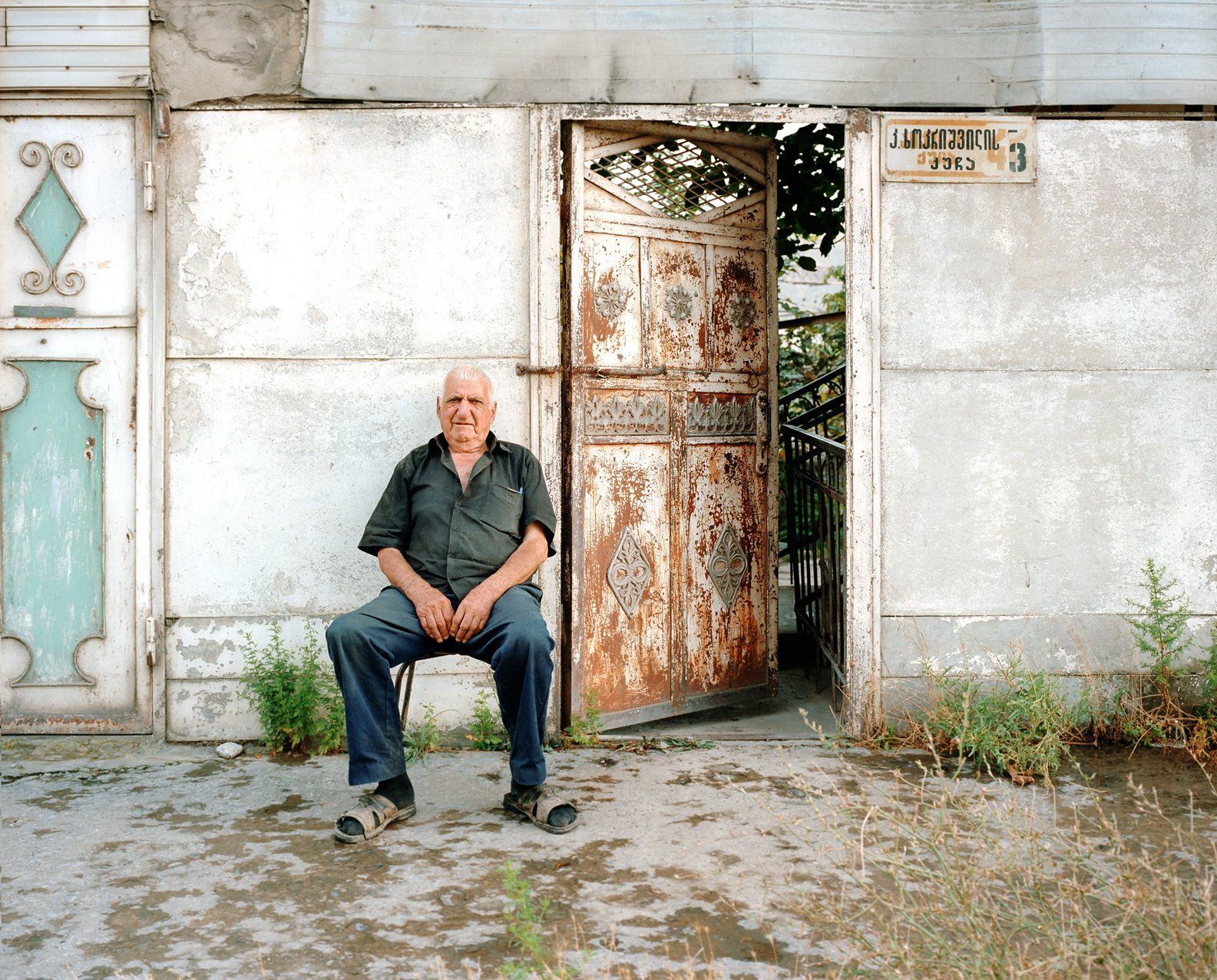 An older man sitting on a chair in front of a dilapidated wall with an open rusty gate, some weeds growing on the ground.