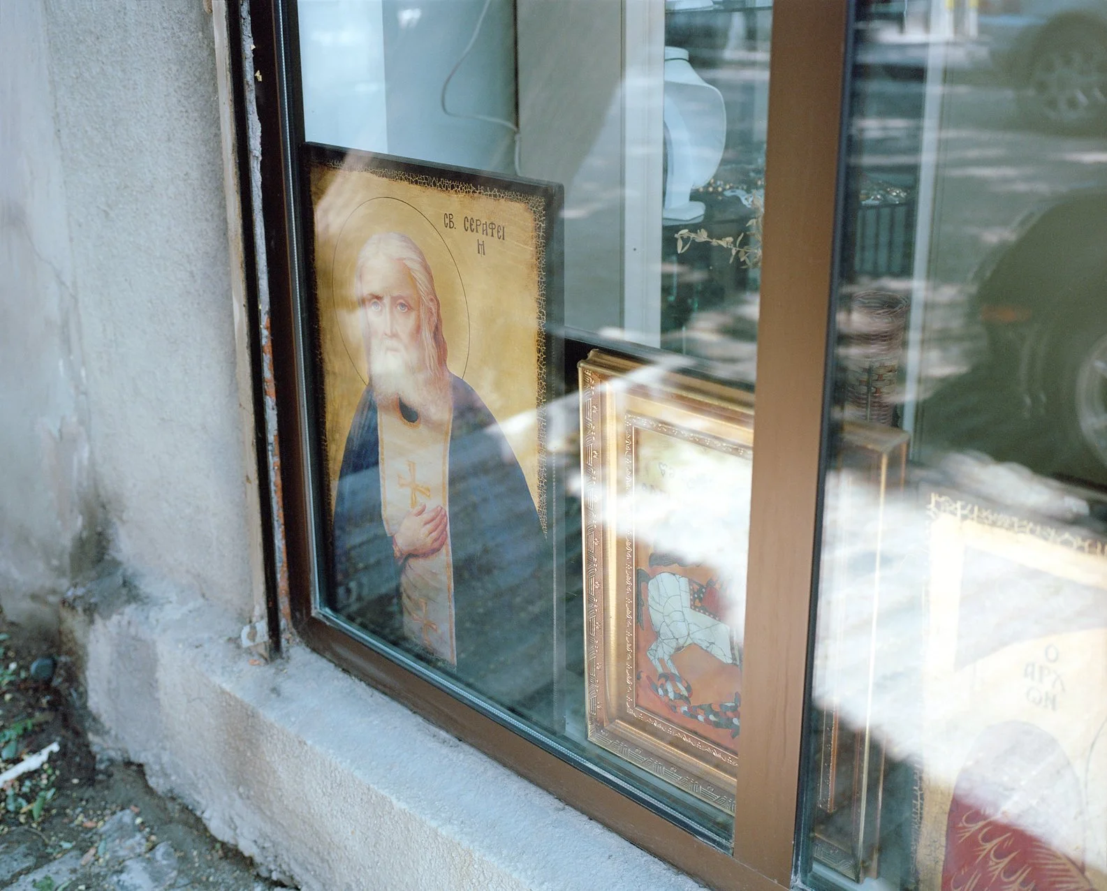 Religious icon of Jesus Christ with a halo, holding a cross, displayed in a church window, with reflections of the street outside.