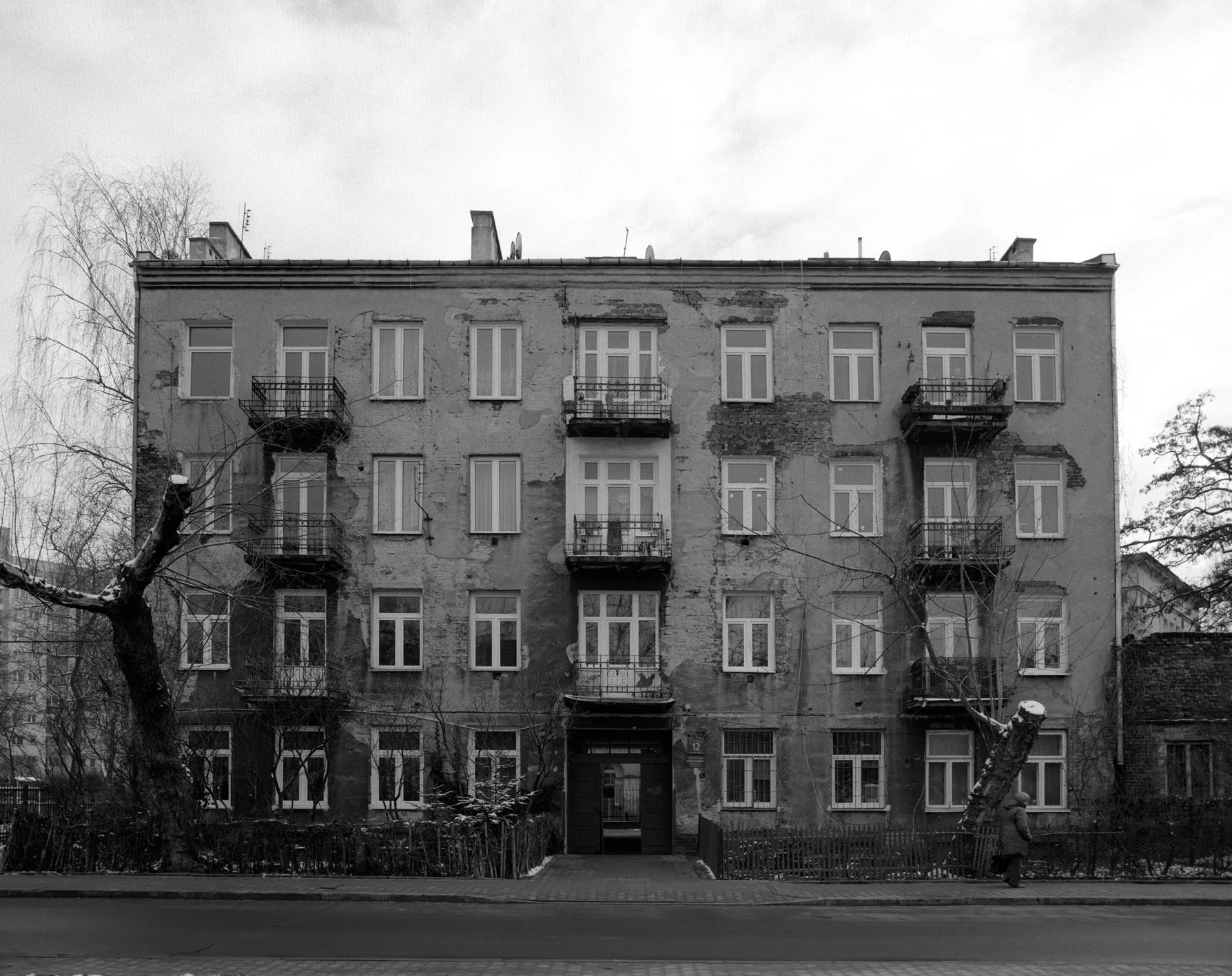 Black and white photo of a weathered, multi-story residential building with peeling paint and cracks, including balconies, windows, and a central entrance, with leafless trees in front and a person walking on the sidewalk.