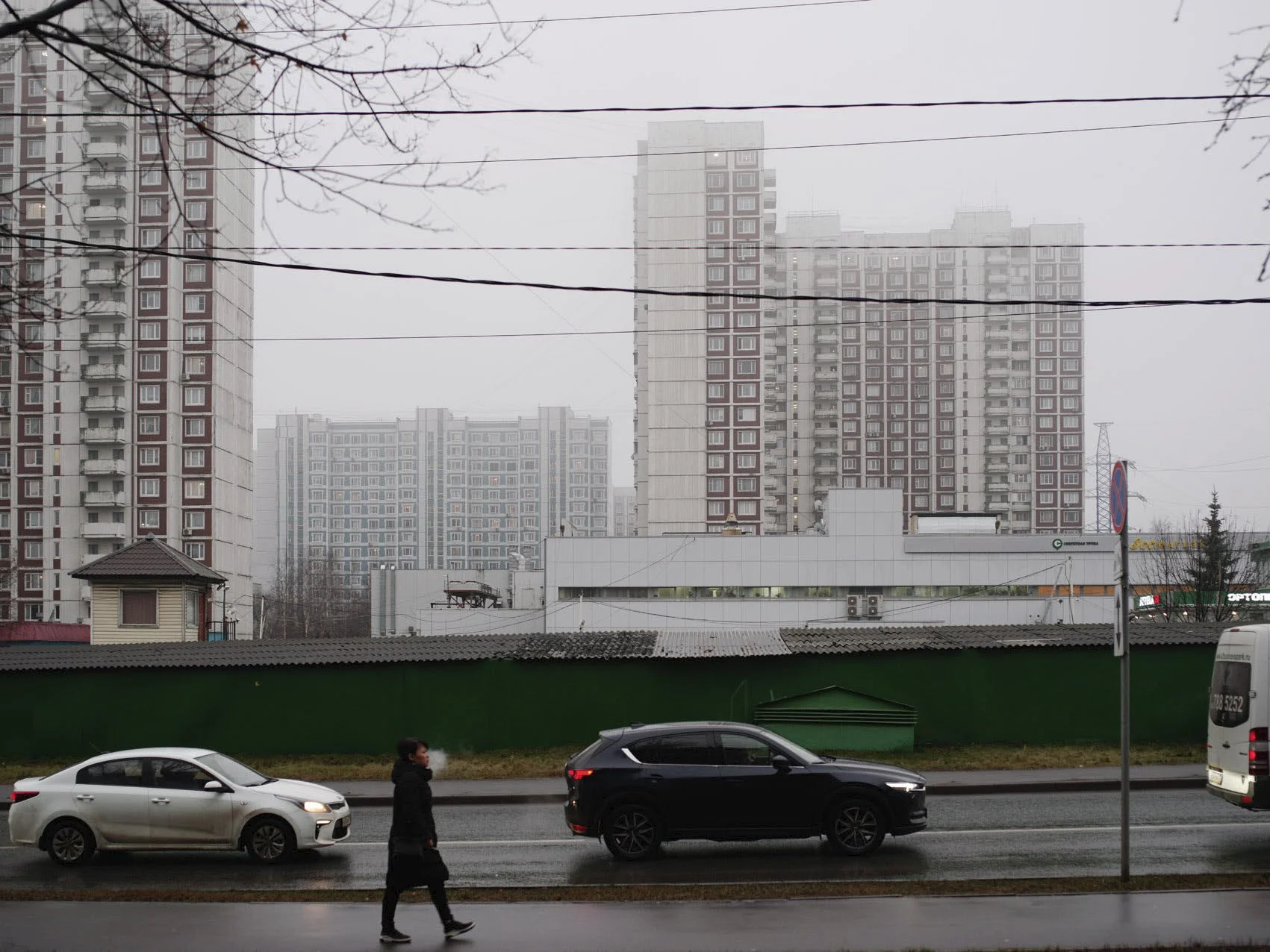 City street scene with a woman walking on wet sidewalk, three parked cars, and high-rise apartment buildings in the foggy background.