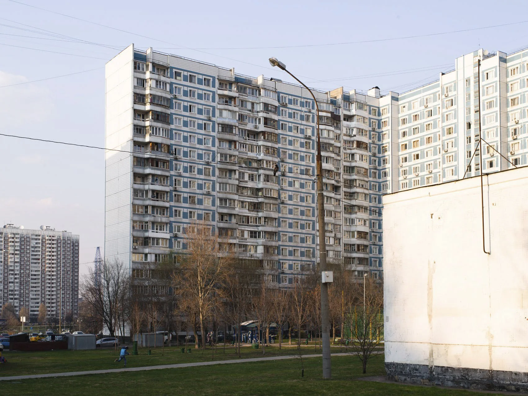 A large apartment building with multiple balconies, a park with trees and children playing, and a white building in front.