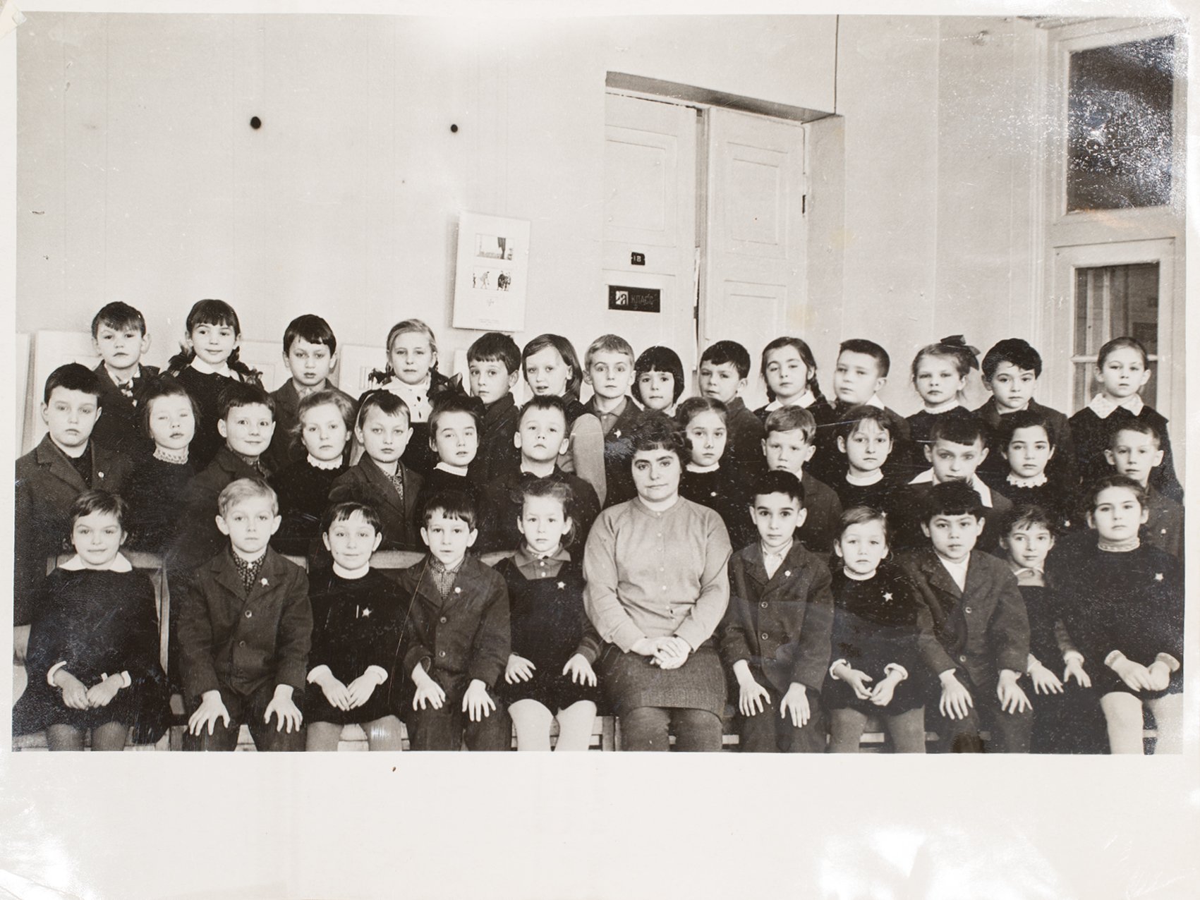 A black and white classroom photo of a school class with 27 children and one adult female teacher, all posed in rows inside the classroom.