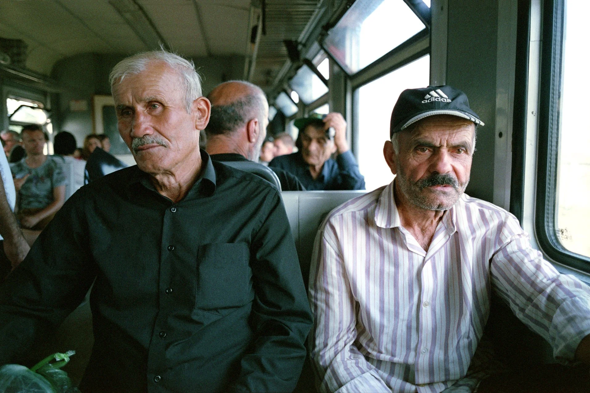 Two elderly men sitting on a train, with other passengers visible in the background. One man wears a black shirt, and the other wears a striped shirt and a black cap.
