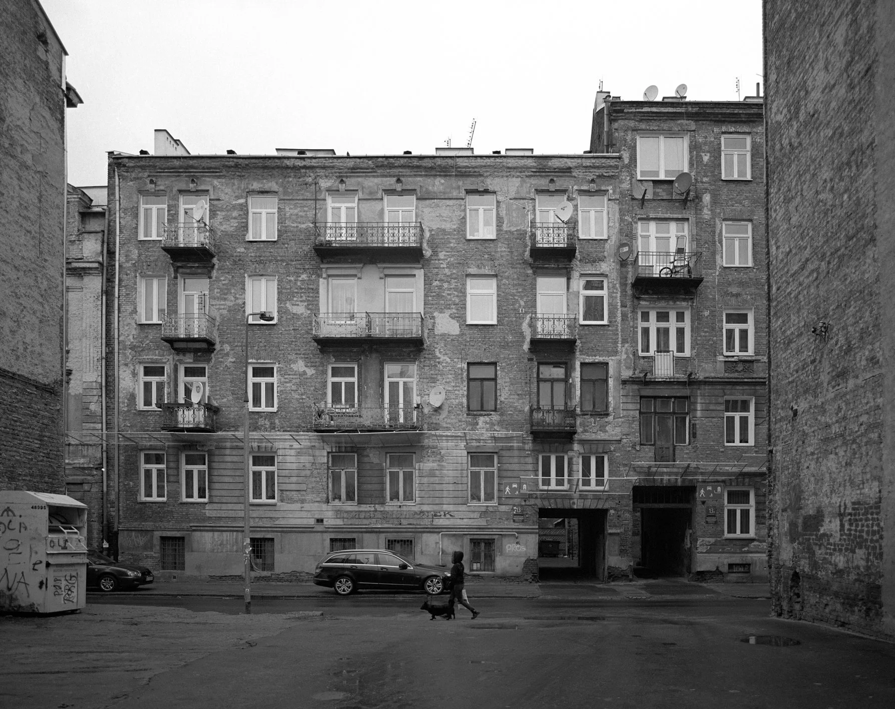 Black and white image of a multi-story apartment building with balconies, satellite dishes, and a street scene with a person walking a dog and parked cars.
