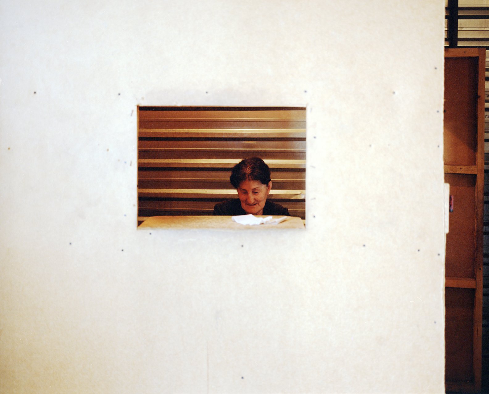 A woman with short dark hair, wearing a black blazer, is seen through a small rectangular cutout in a large beige partition or wall. She appears to be reading or looking down at something on a table in front of her, with a striped wooden background behind her.