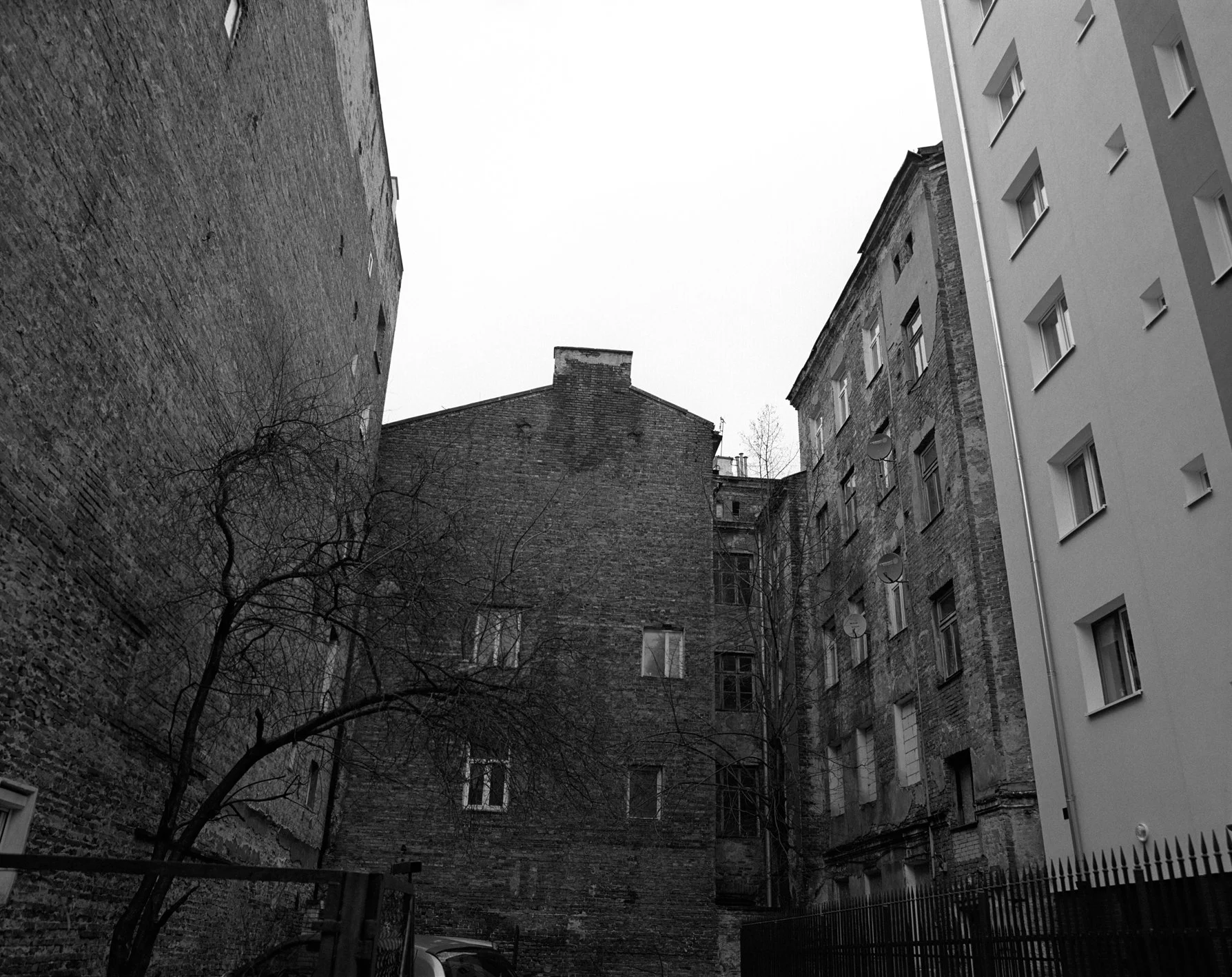 A black and white photo of old brick buildings with multiple windows, some with satellite dishes, and a leafless tree in the foreground.