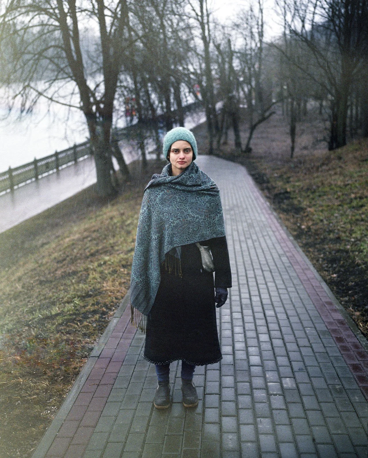 A woman dressed warmly in a gray beanie, a patterned shawl, a long black skirt, and boots stands on a paved pathway in a park on a cold, overcast day. Bare trees and a fence are visible in the background.