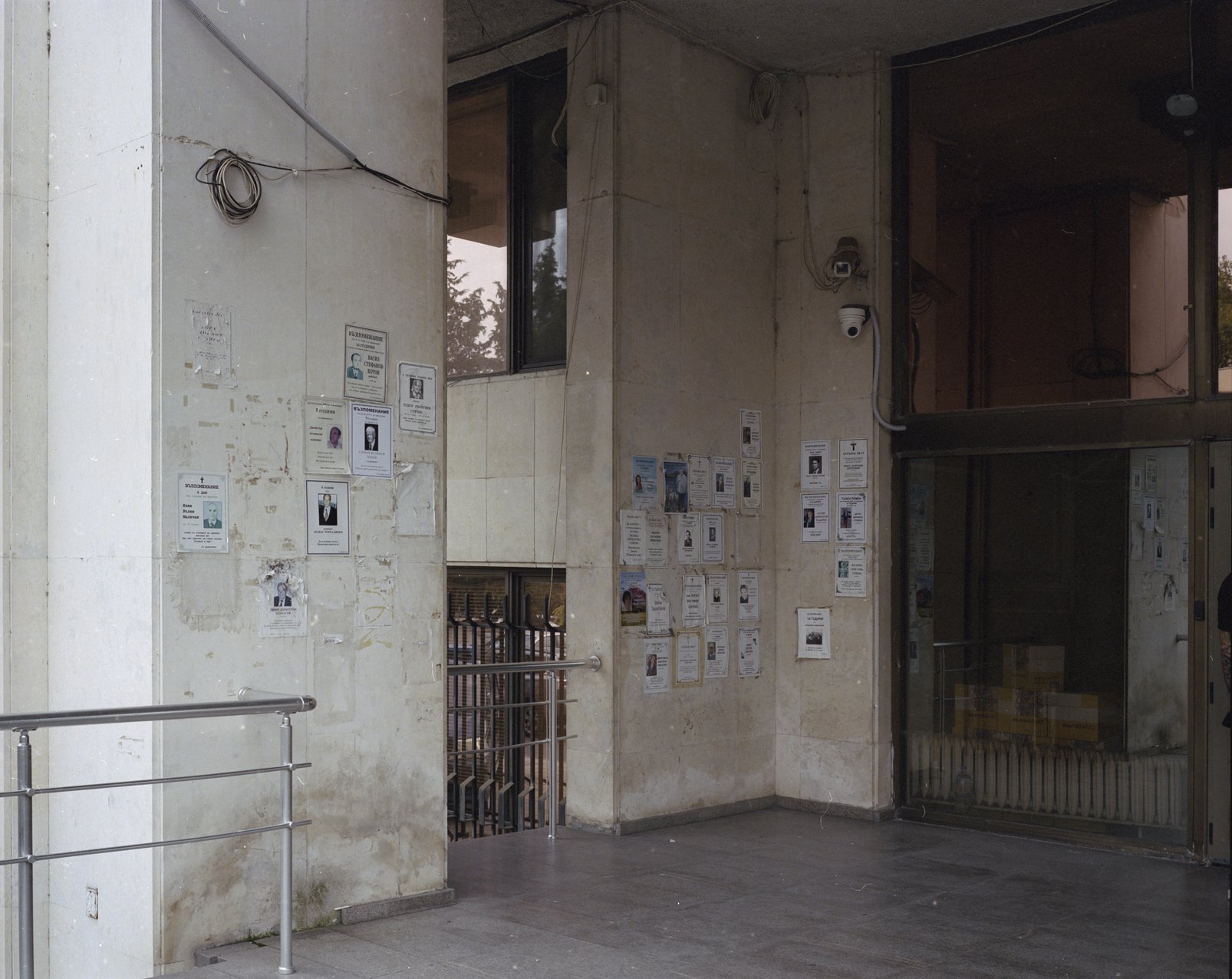 A building entrance with multiple missing persons and wanted posters on concrete walls, security cameras, and a glass door.