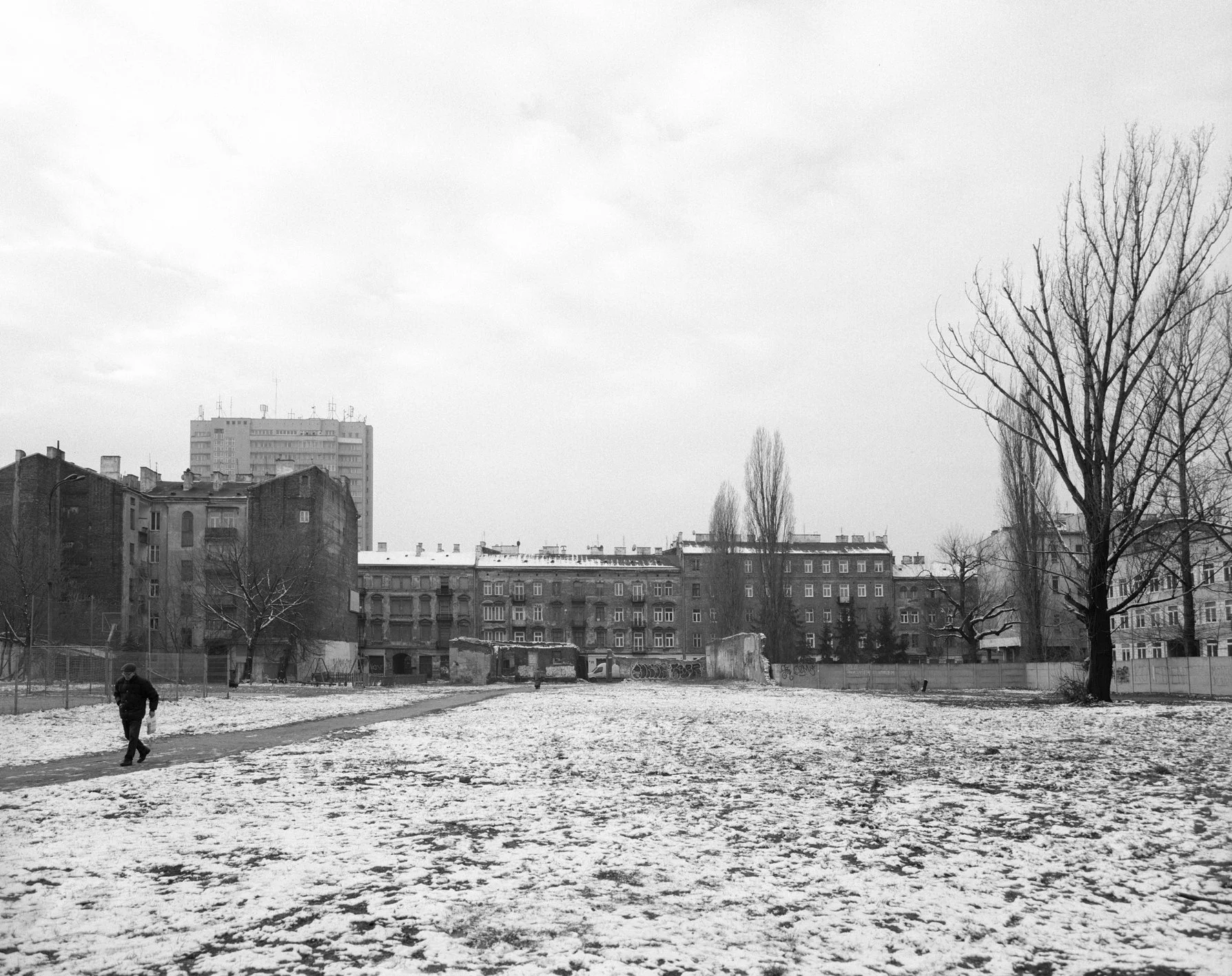 A person walking on a snow-covered field with leafless trees and old apartment buildings in the background, overcast sky.