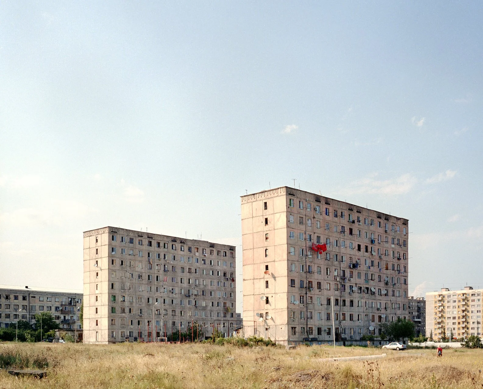 Two large apartment buildings with multiple stories and many windows, set in an open grassy area with a few trees and a person riding a bicycle in the foreground, under a partly cloudy sky.