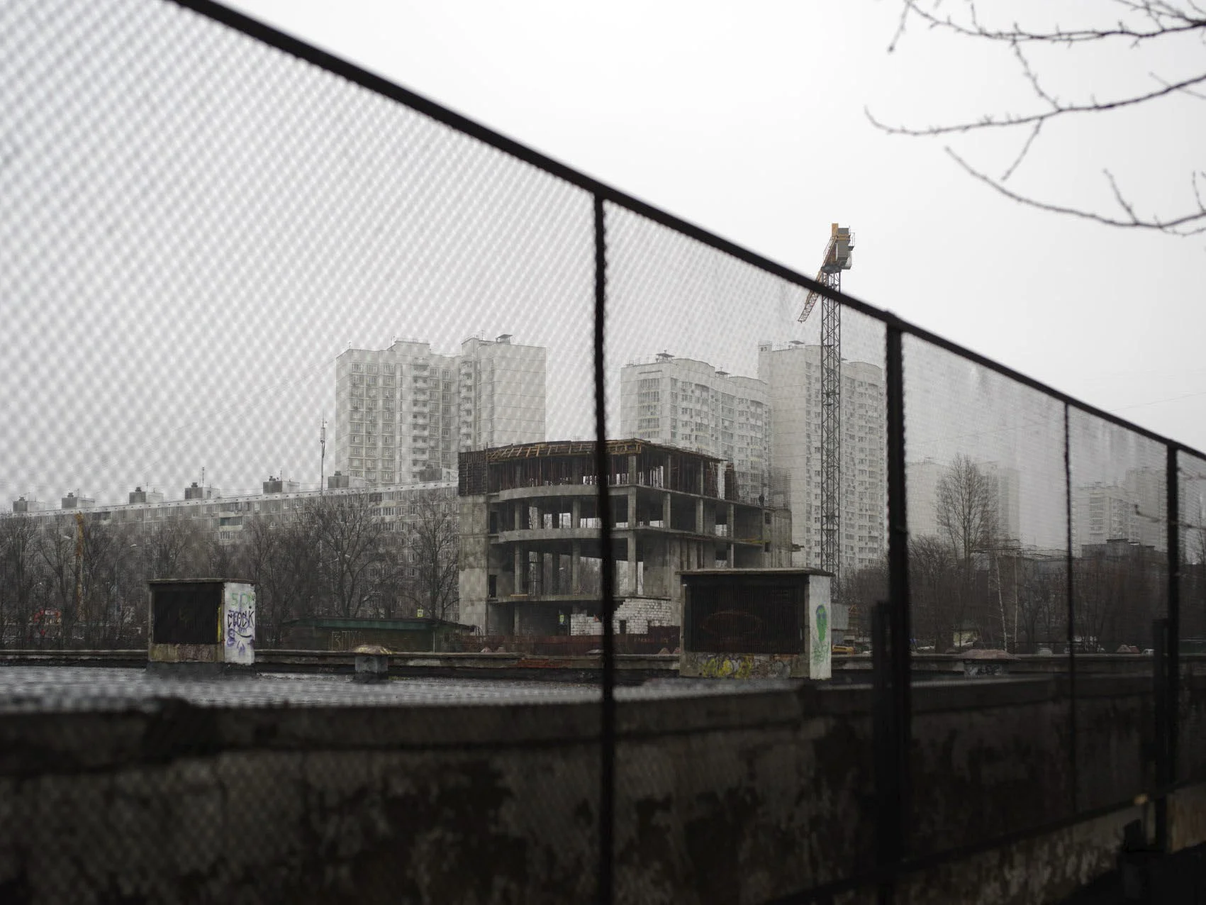 View of a cityscape seen through a chain-link fence, with a construction site, unfinished building, high-rise apartment buildings, a crane, and overcast sky in the background.